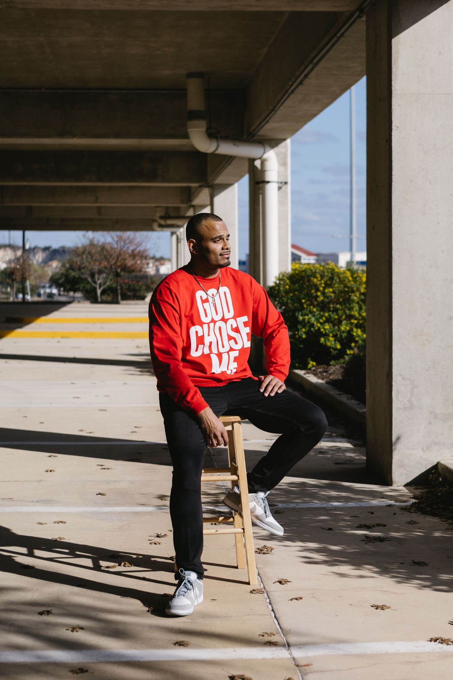 Man in red God Chose Me sweatshirt sitting on stool under concrete structure outdoors.