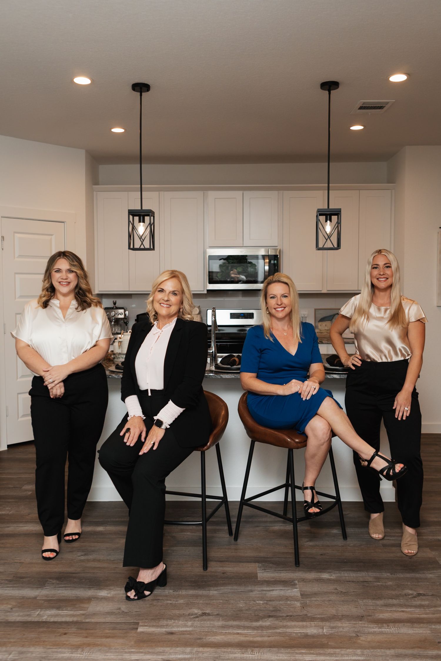 Four women in business attire pose together in a modern kitchen with pendant lights and white cabinets.