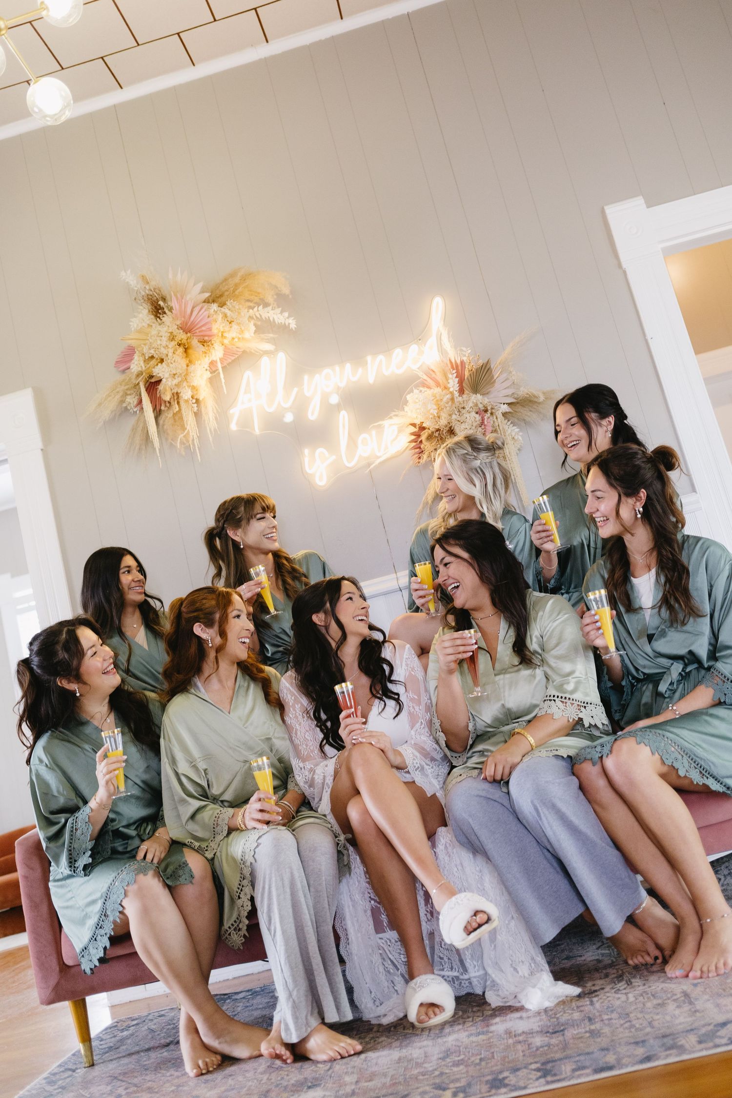 Bridesmaids in sage and gray robes laughing together on a couch, holding mimosas under an All You Need Is Love neon sign.