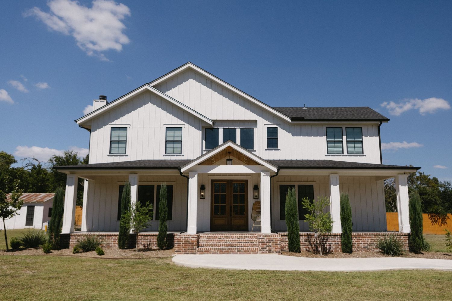 Modern white farmhouse with covered porch, brick accents, black trim windows, and lush green lawn under blue sky.