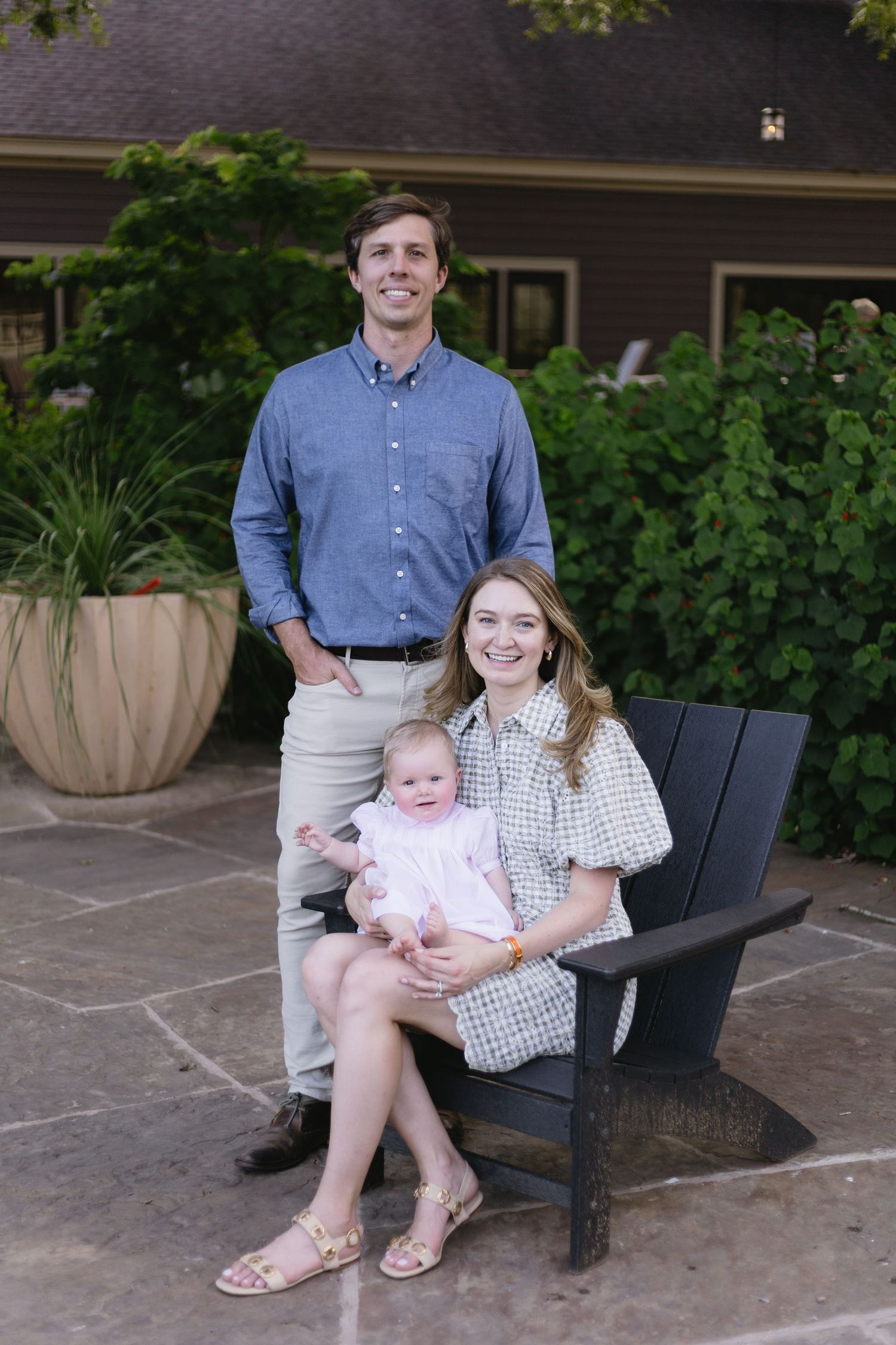 Young family portrait with man standing behind woman seated in Adirondack chair holding baby in pink dress outdoors.
