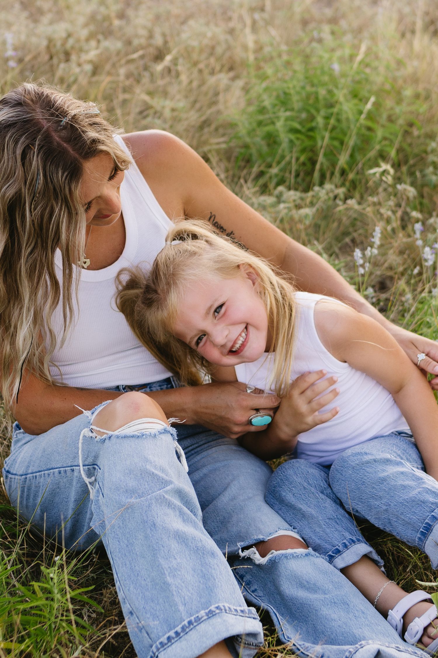 Mother and daughter in matching white tanks and jeans laughing together in a sunny field.