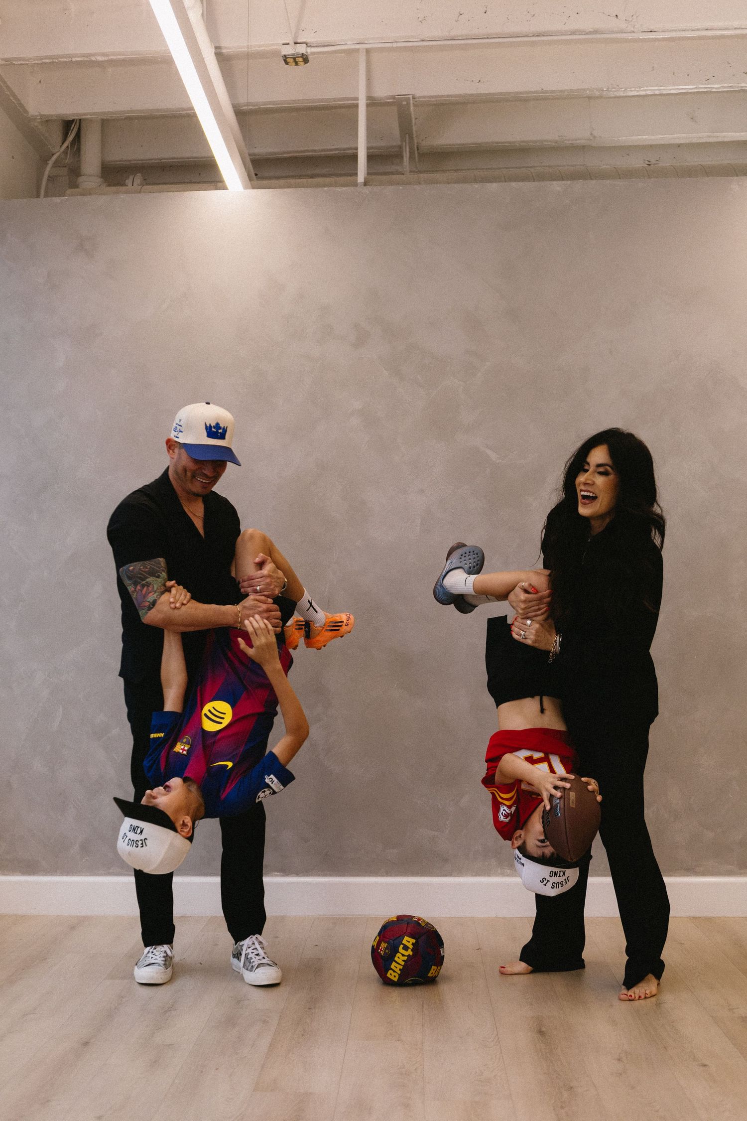 A couple playfully holds two children upside down in a modern studio with a soccer ball on the floor.