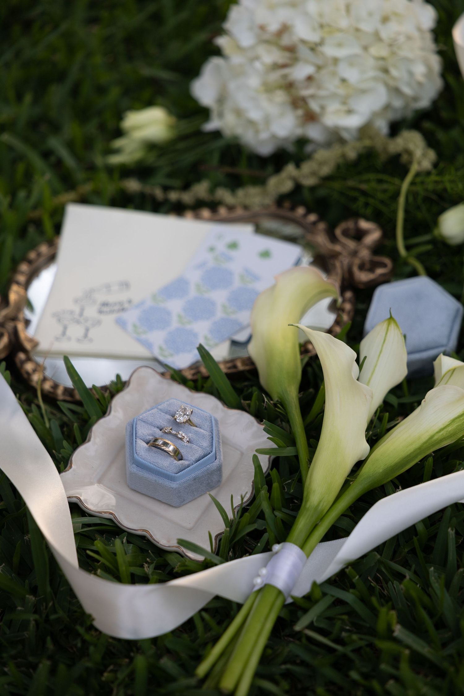 Wedding rings in a blue velvet box beside calla lilies, white hydrangeas, and stationery on green grass.