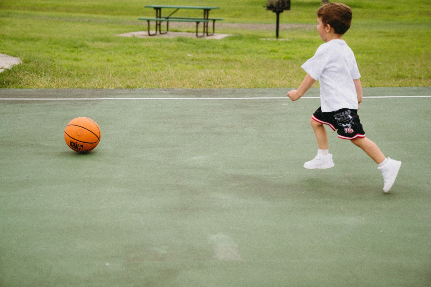 Young boy in white shirt and black shorts chasing an orange basketball on an outdoor court.