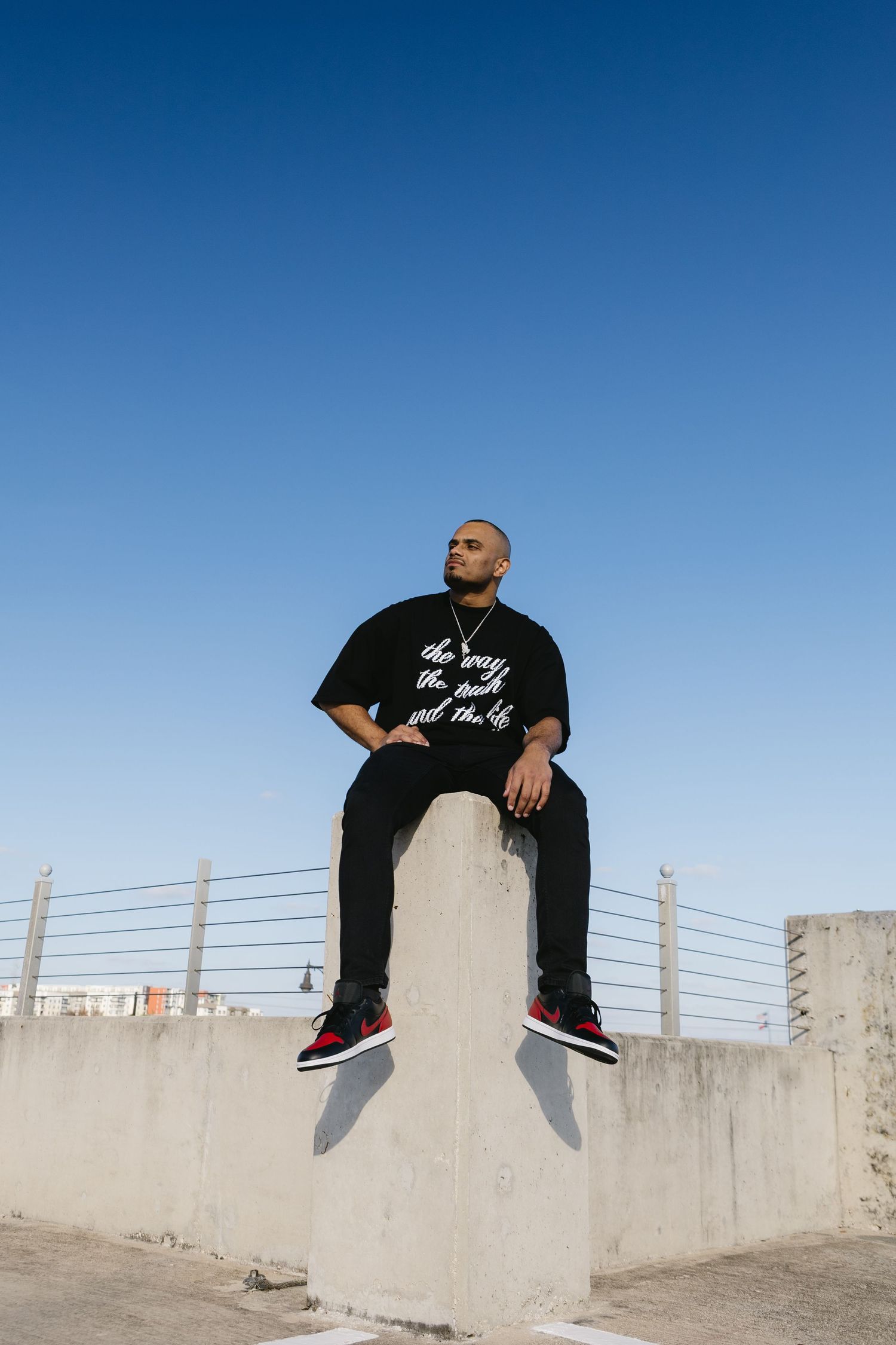 Man in black graphic tee sitting on concrete wall against clear blue sky wearing Jordan sneakers.