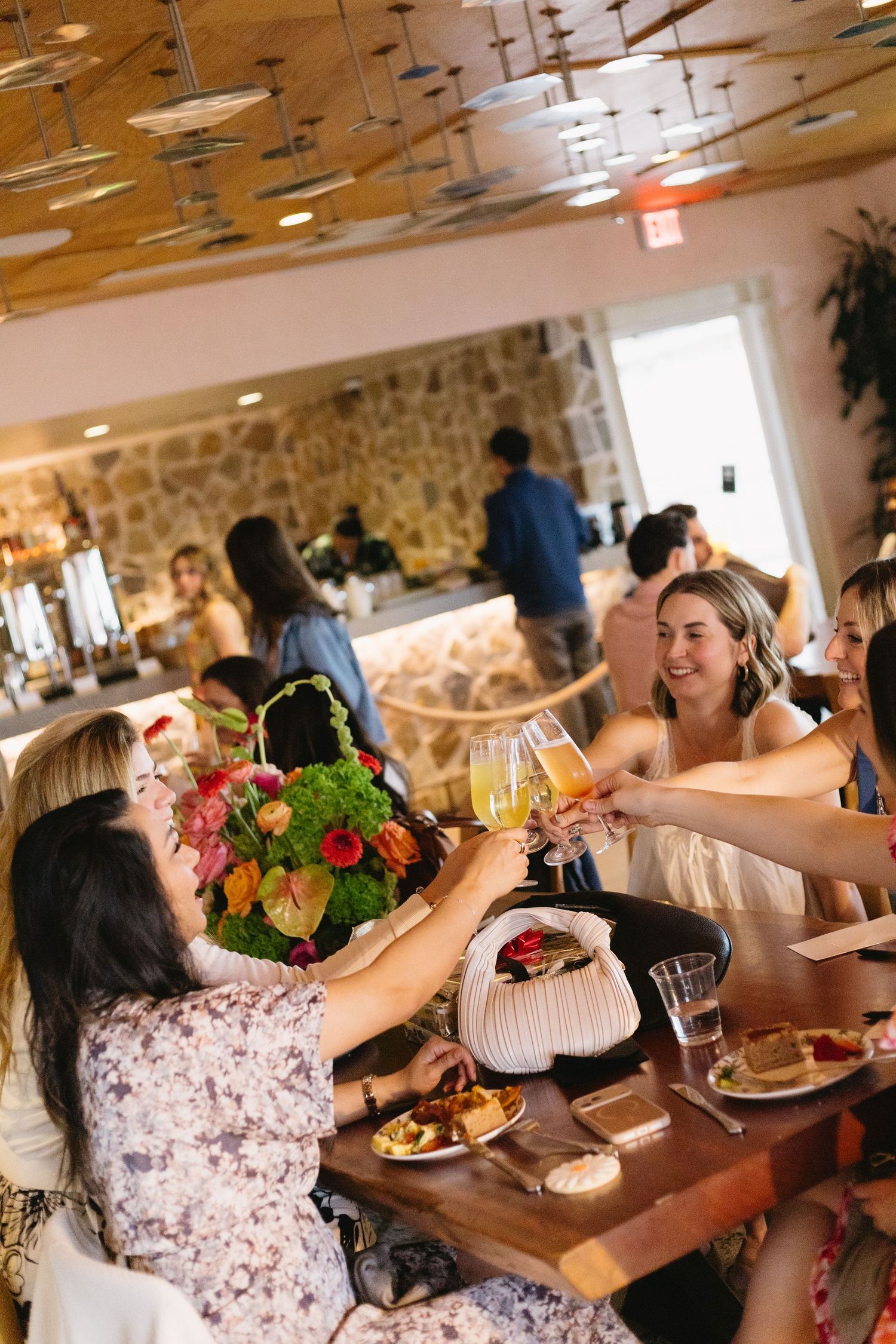 Group of women toasting with champagne glasses over brunch at a stylish restaurant with floral centerpiece.