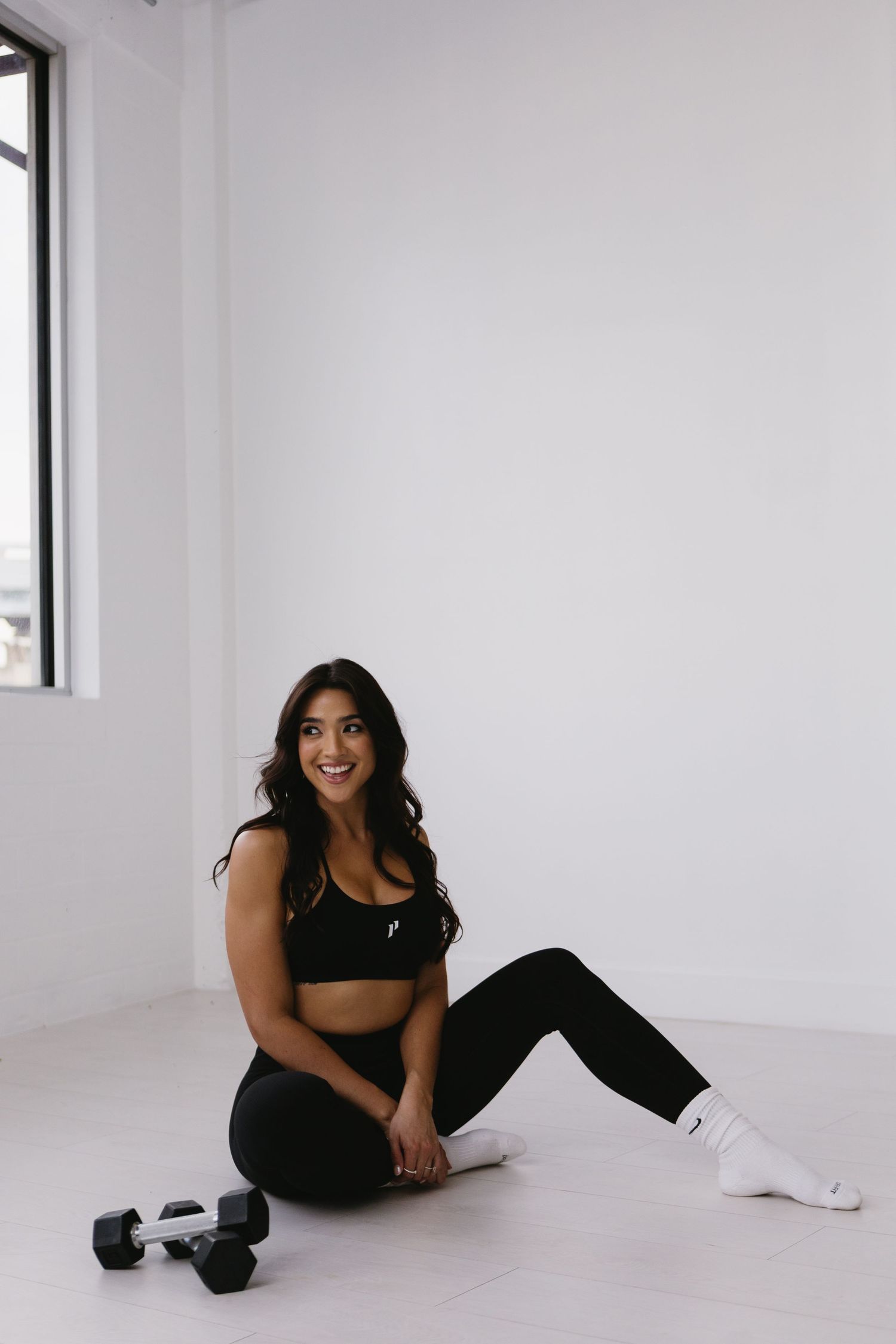 Woman in black sports bra and leggings sitting on floor with dumbbells, smiling in bright white studio.