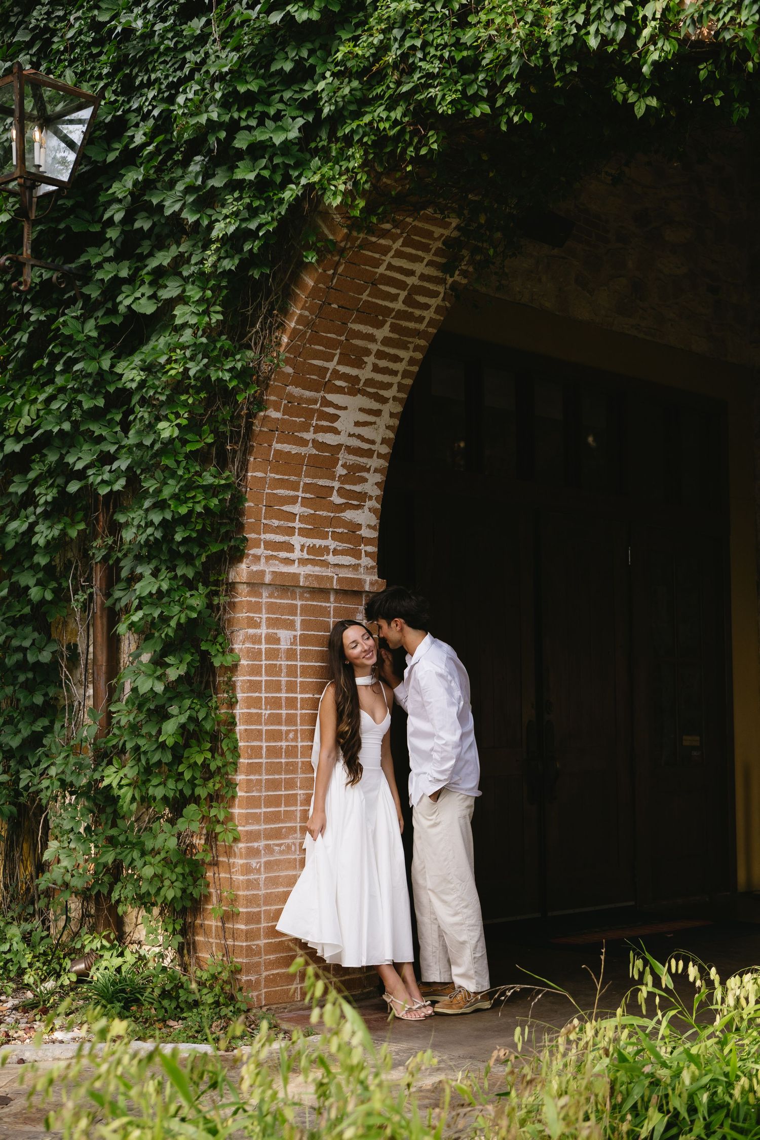 Couple sharing intimate moment under ivy-covered brick archway surrounded by lush greenery.