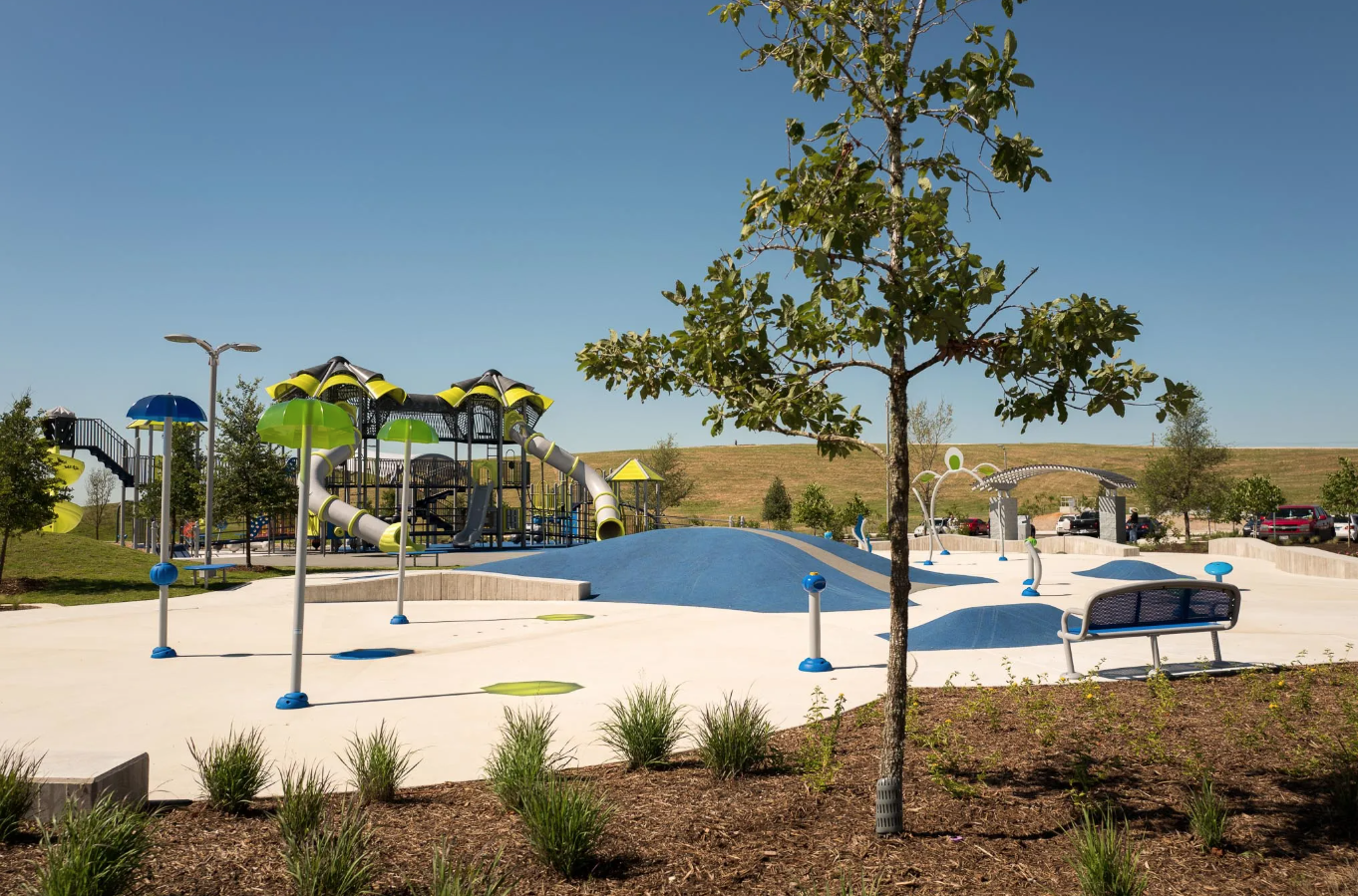 Modern outdoor splash pad and playground area with colorful play structures, water features, and landscaping under a clear blue sky.