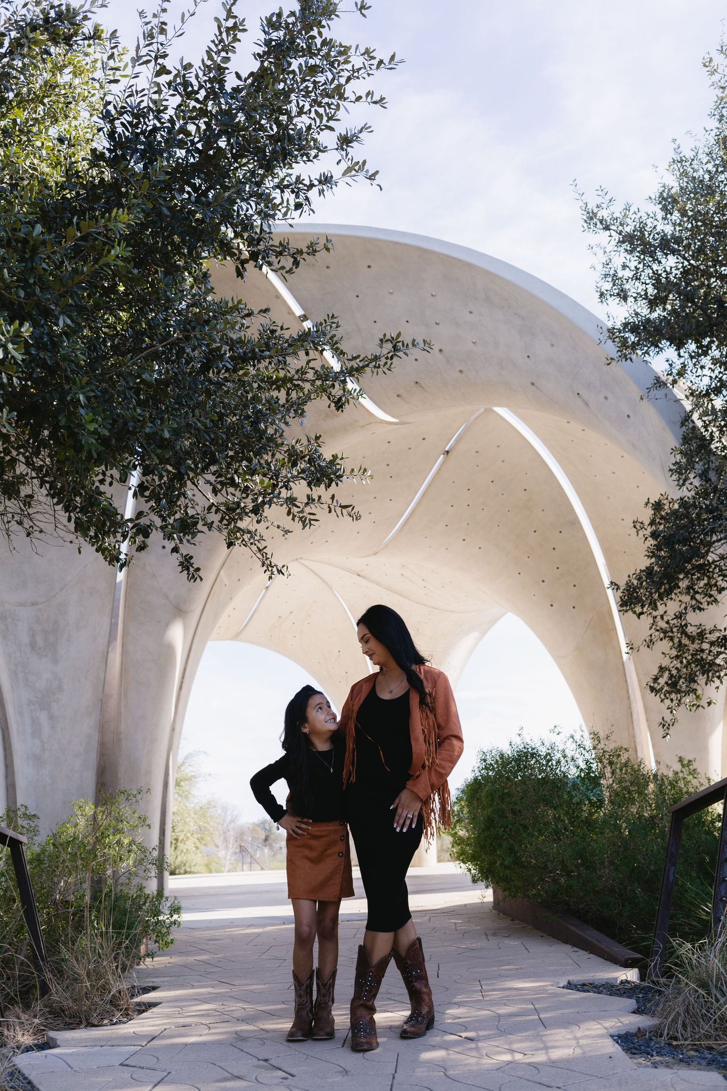 A woman and young girl in matching brown skirts and boots stand together under a modern arch structure outdoors.