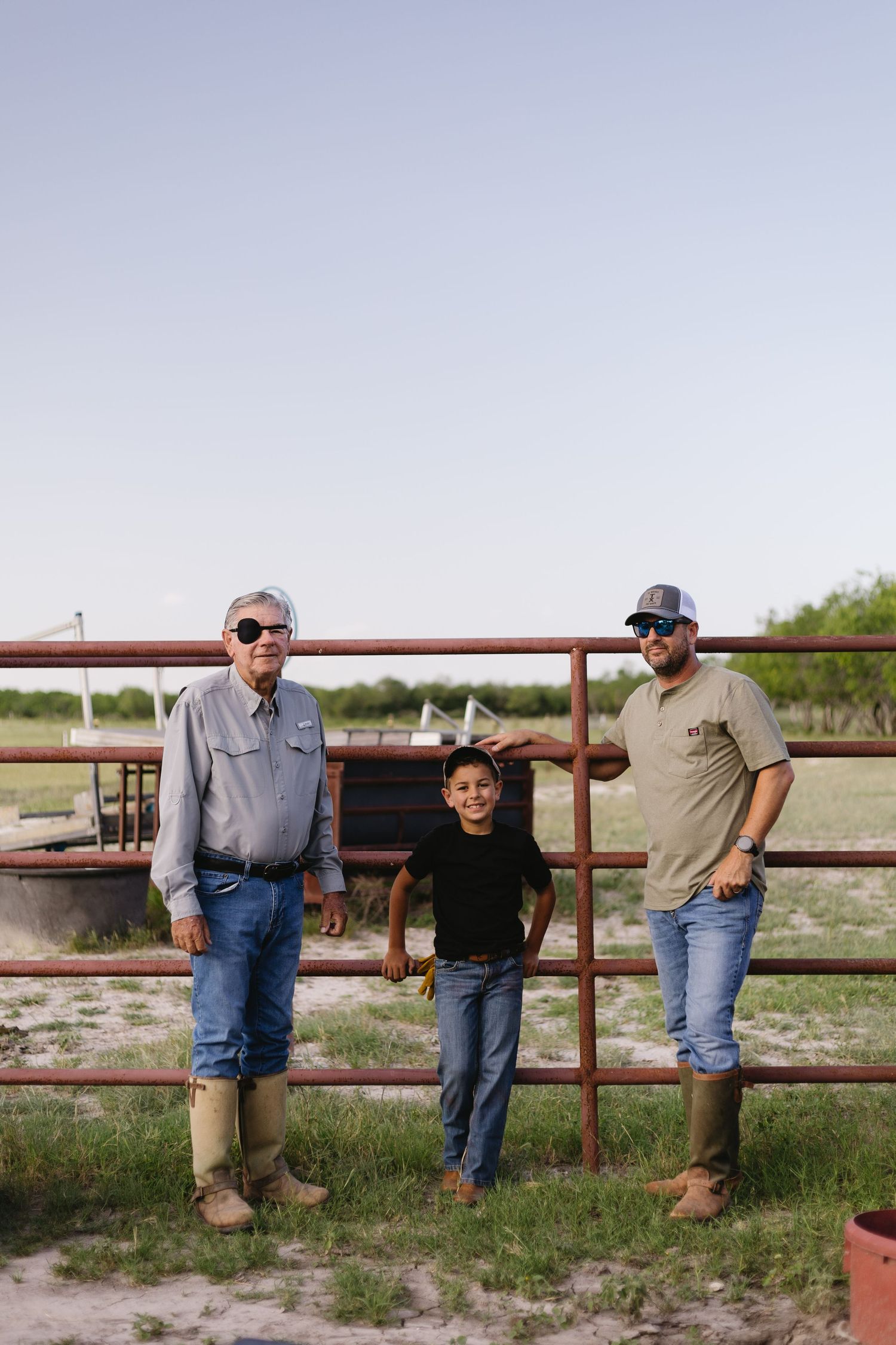 Three generations pose together leaning against a rustic metal gate on a rural farm.