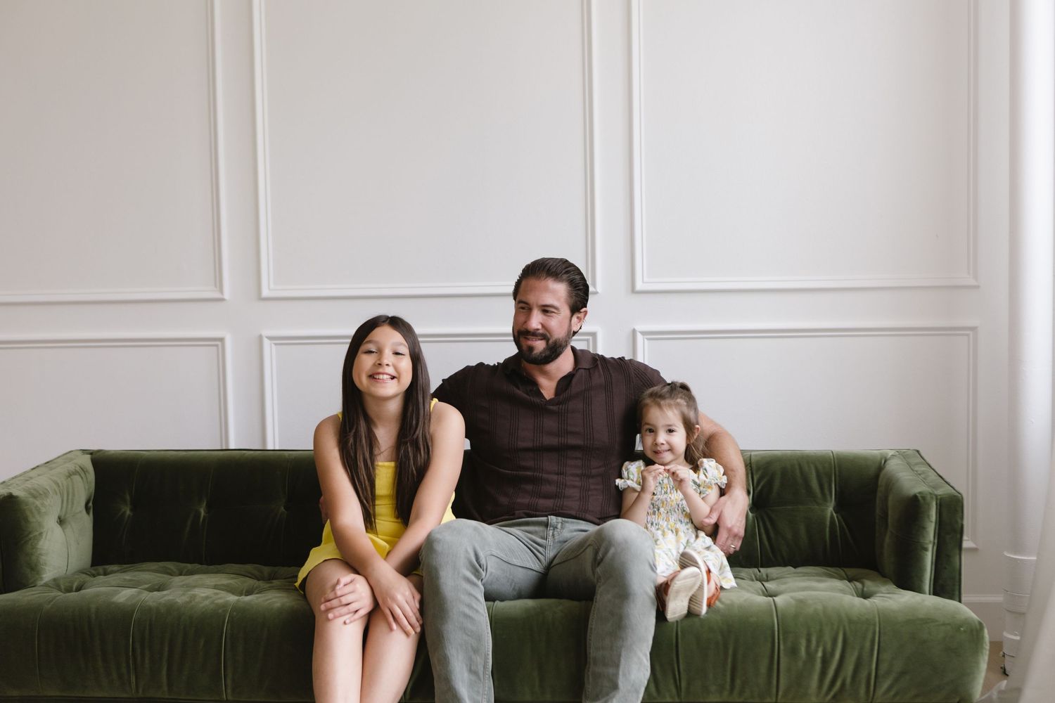 A family relaxes together on a plush green velvet sofa in a bright white room with wainscoting.