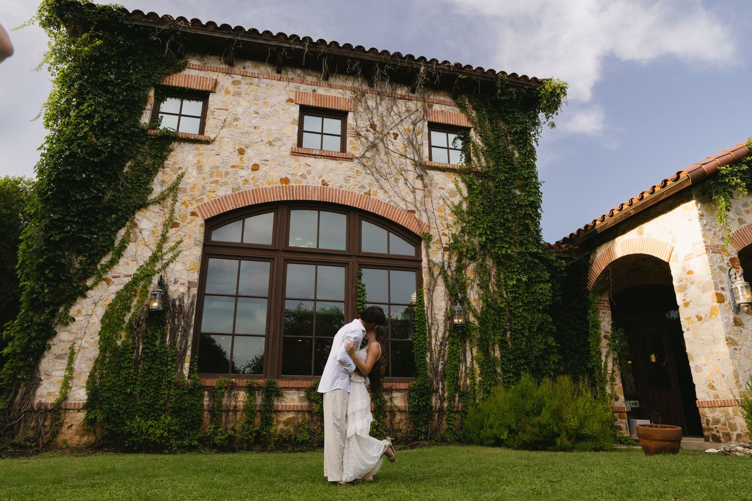 Couple sharing a romantic kiss on green lawn outside ivy-covered stone villa wedding venue.