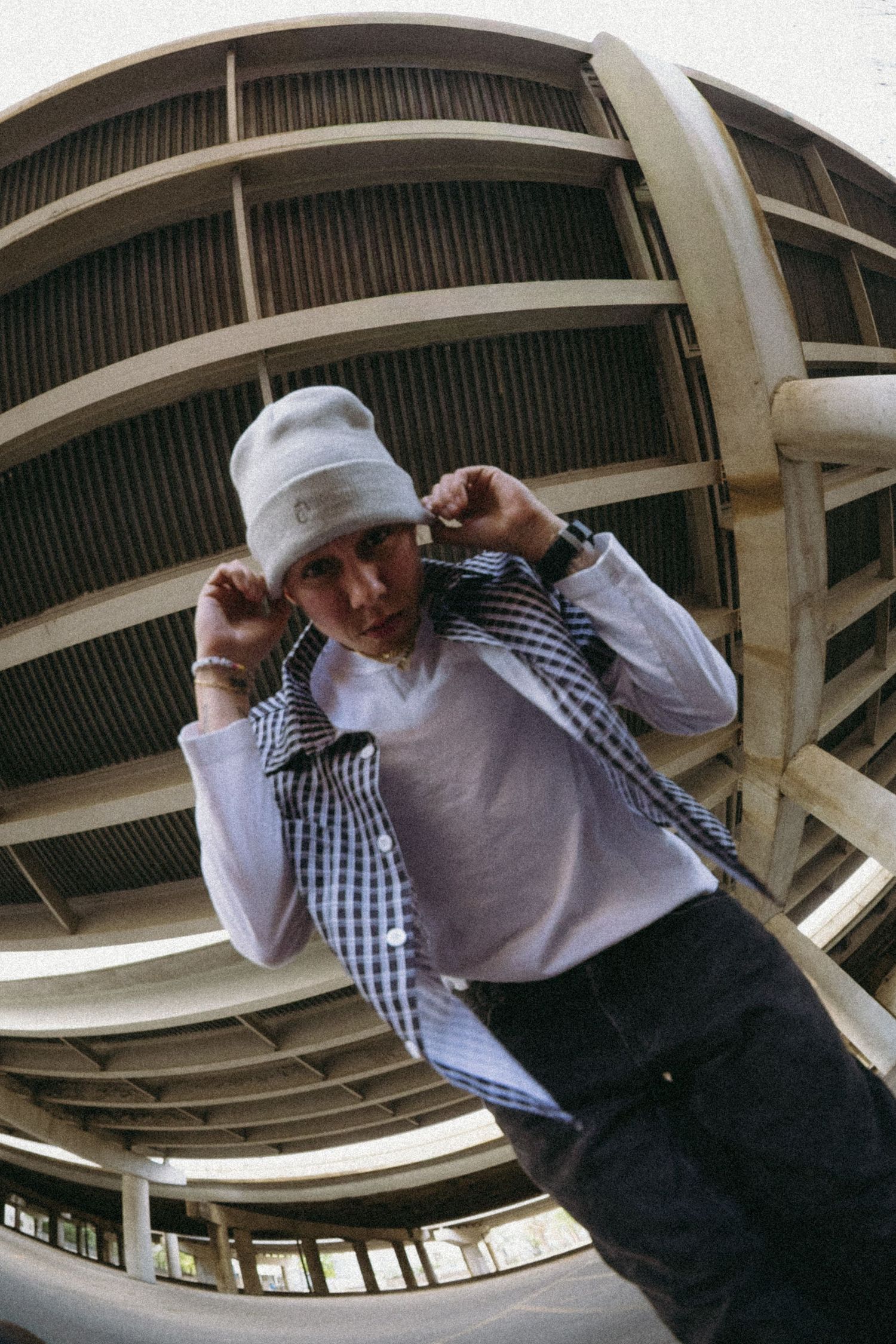 Young man in white beanie and plaid shirt poses under curved parking garage, shot with fisheye lens.