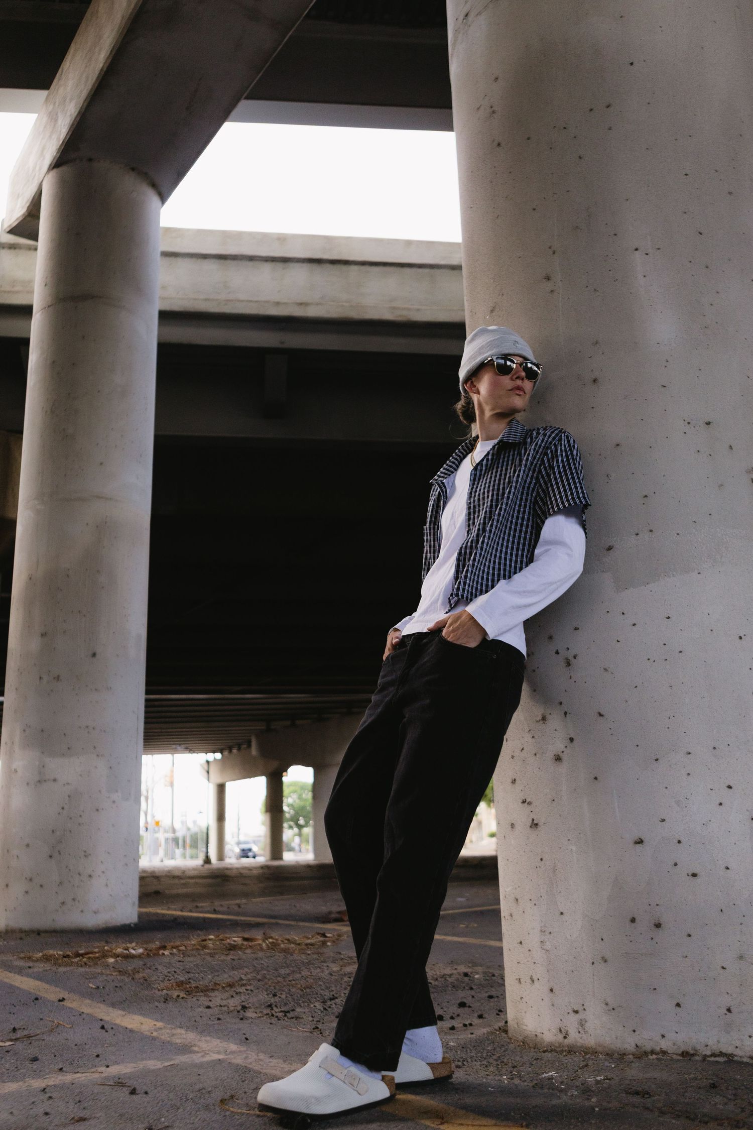 Young man in streetwear leaning against concrete pillar under urban overpass, wearing cap, sunglasses, and white sneakers.