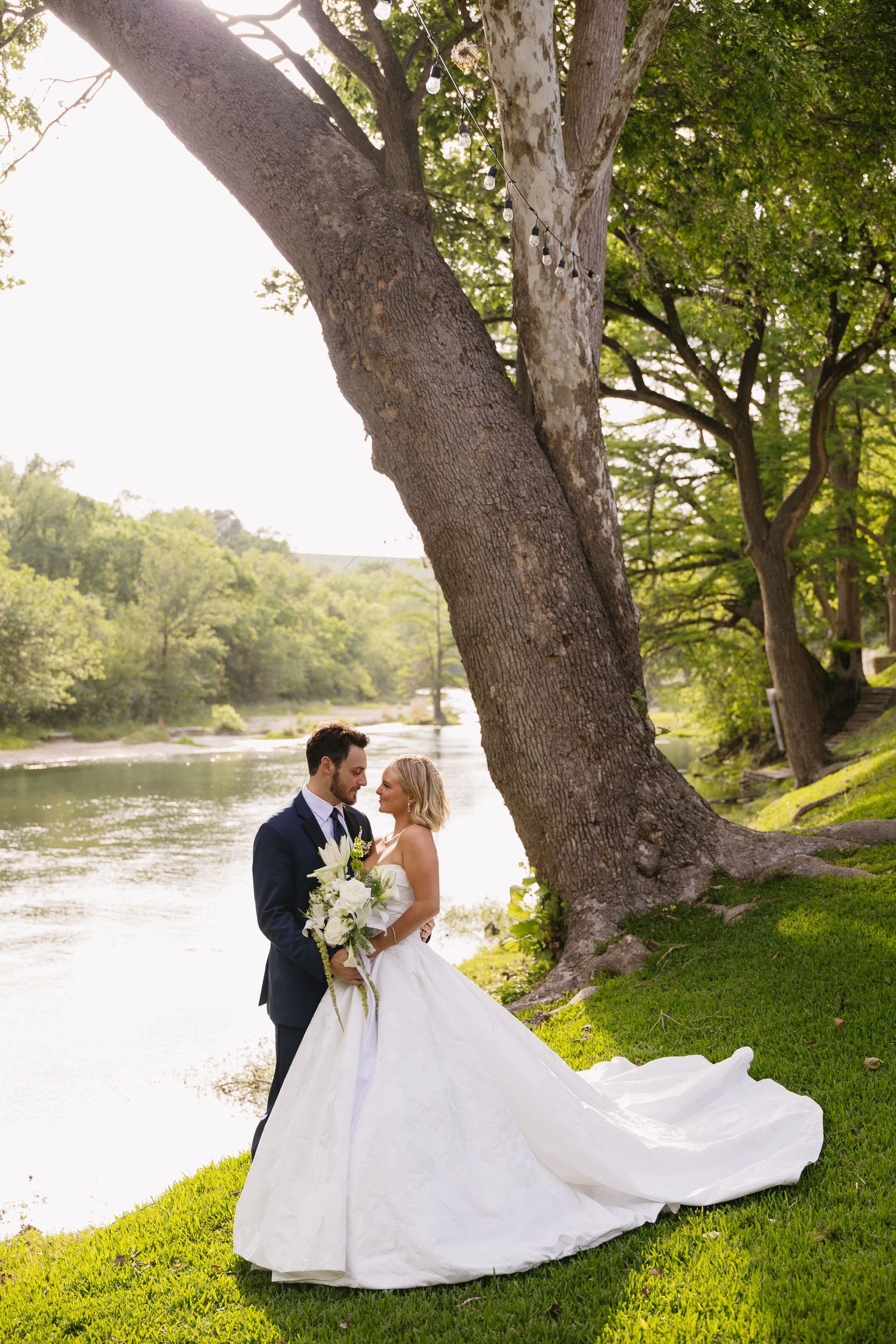 Bride and groom share a romantic moment by a river under a large tree, bride holding white floral bouquet.