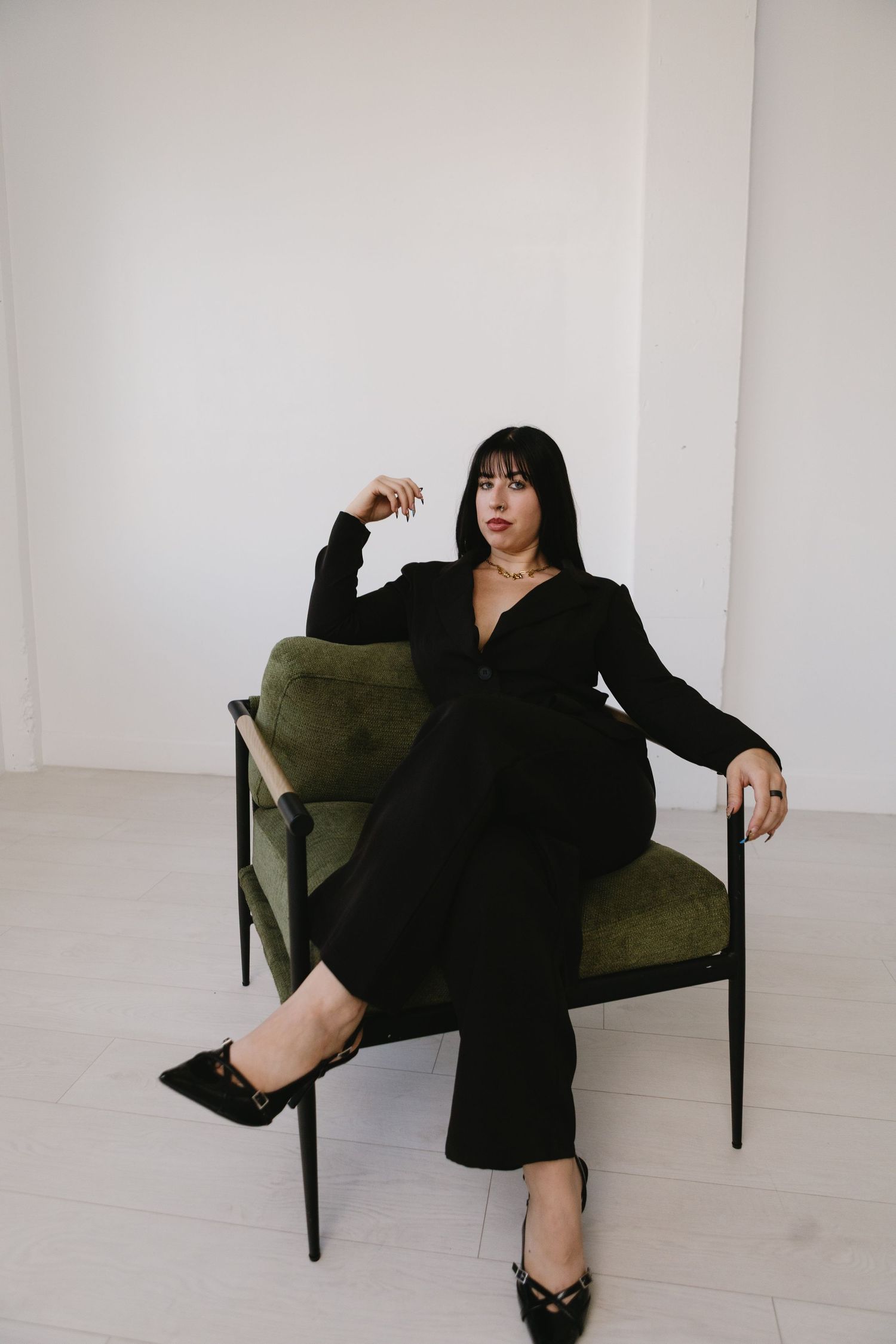 Woman in black jumpsuit and heels seated on green velvet chair against white wall.