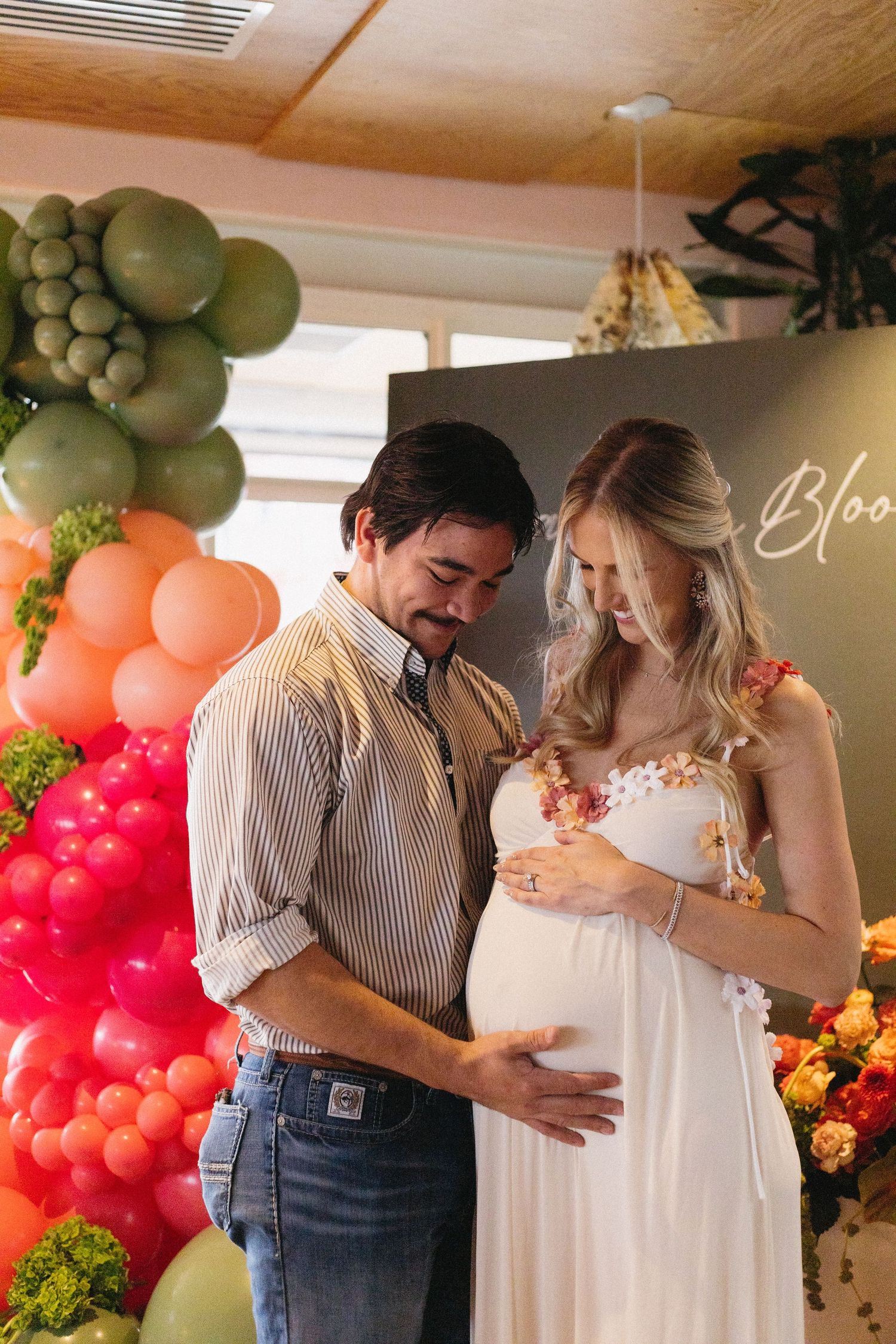 Couple at a baby shower, man touching pregnant woman's belly, colorful balloon backdrop with green, orange, and pink balloons.