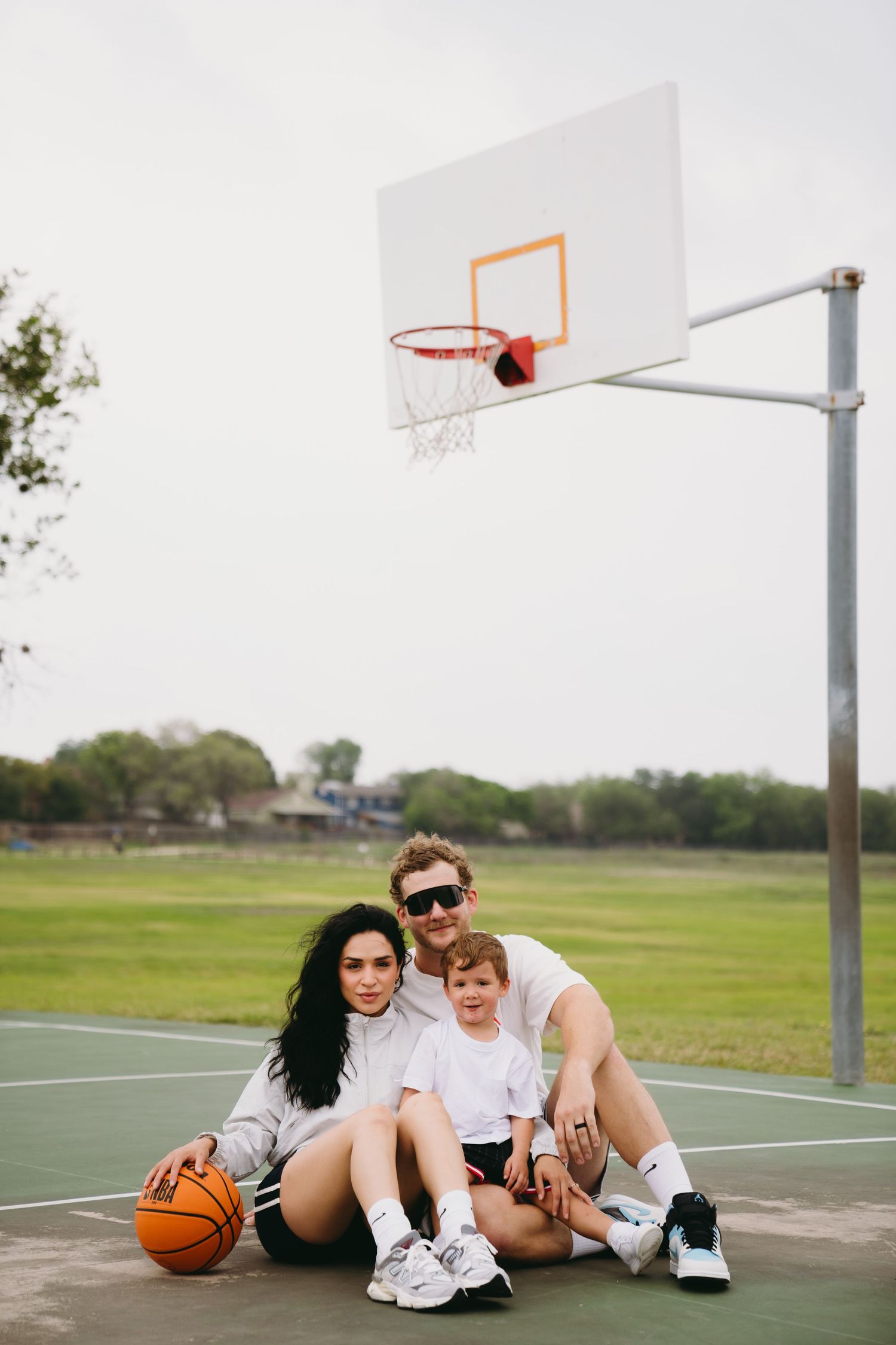 Family of three sitting on outdoor basketball court with hoop, holding orange basketball, wearing white shirts.