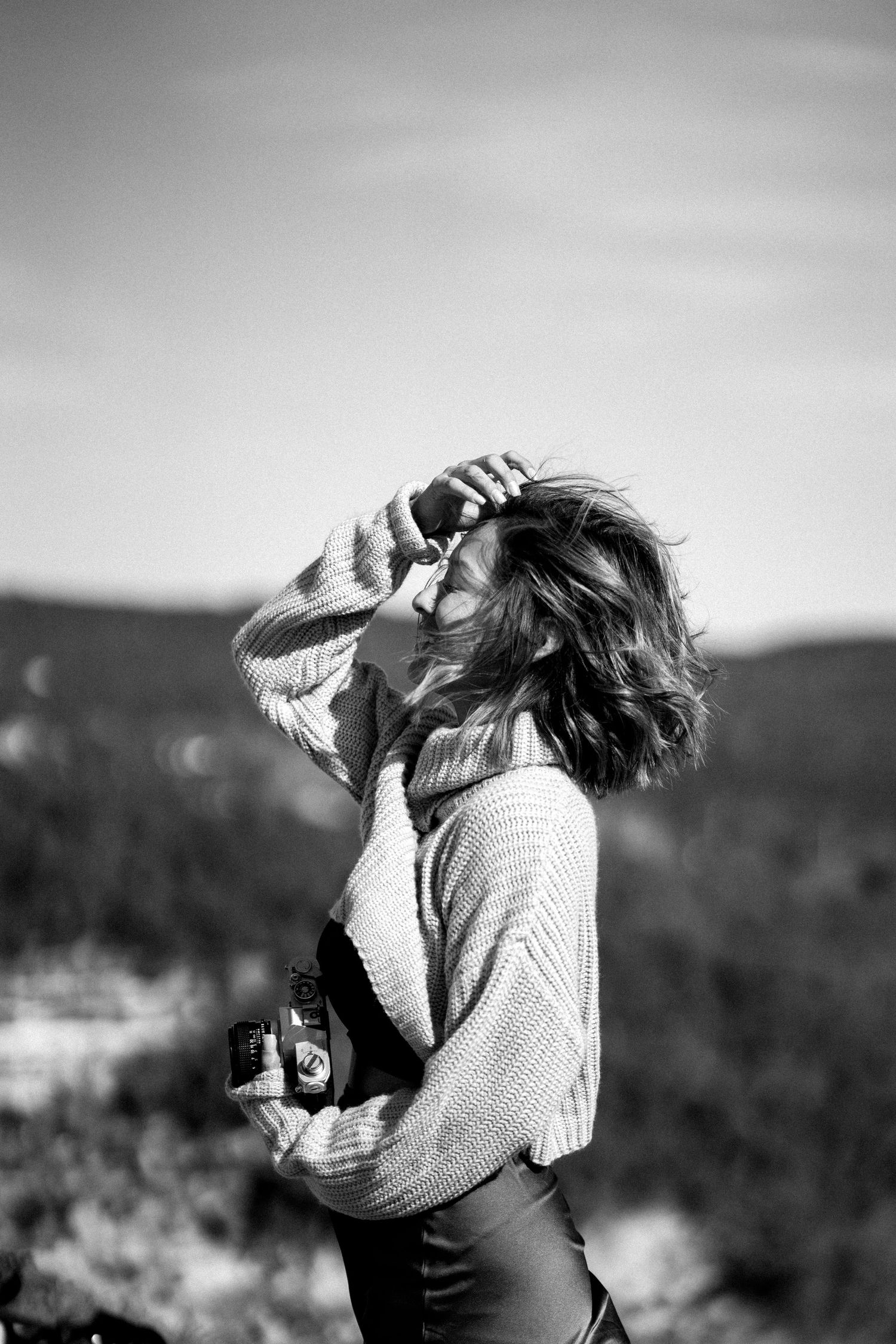 Black and white photo of a woman holding a camera outdoors, shielding her eyes from the sun with mountains behind.