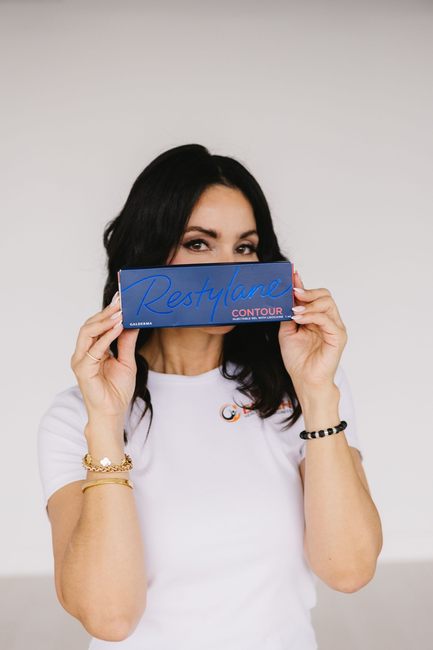 Woman in white t-shirt holding a blue Restylane Contour dermal filler box in front of her face.