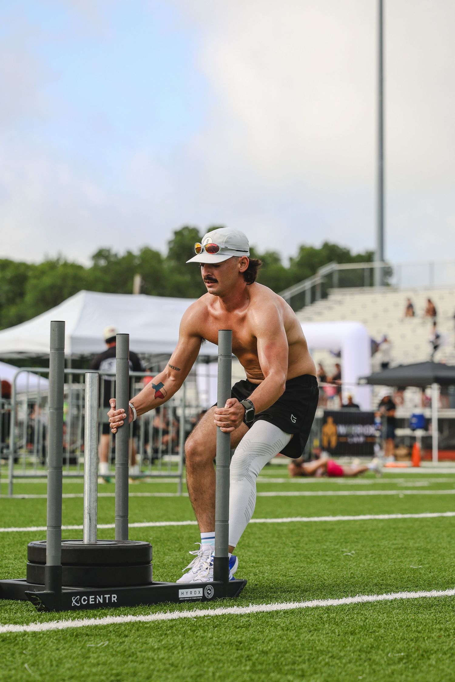Shirtless athletic man pushing weighted sled on turf field during outdoor fitness competition.