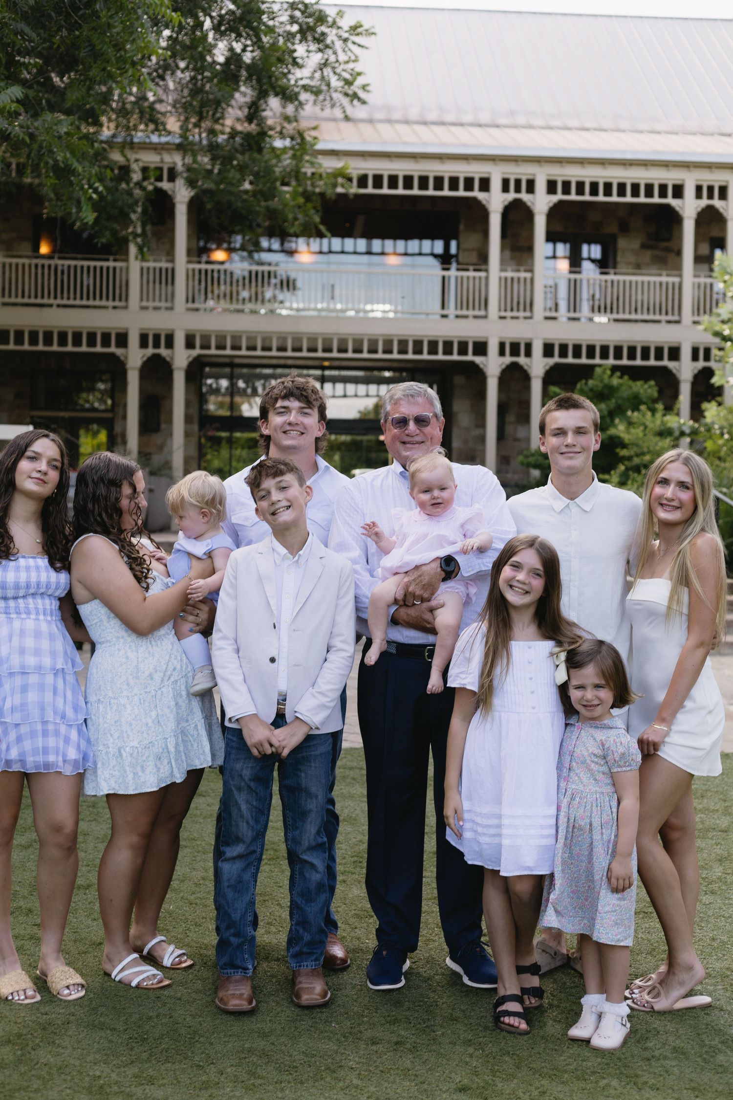 Large multi-generational family group posing outdoors on green lawn in front of elegant two-story building.