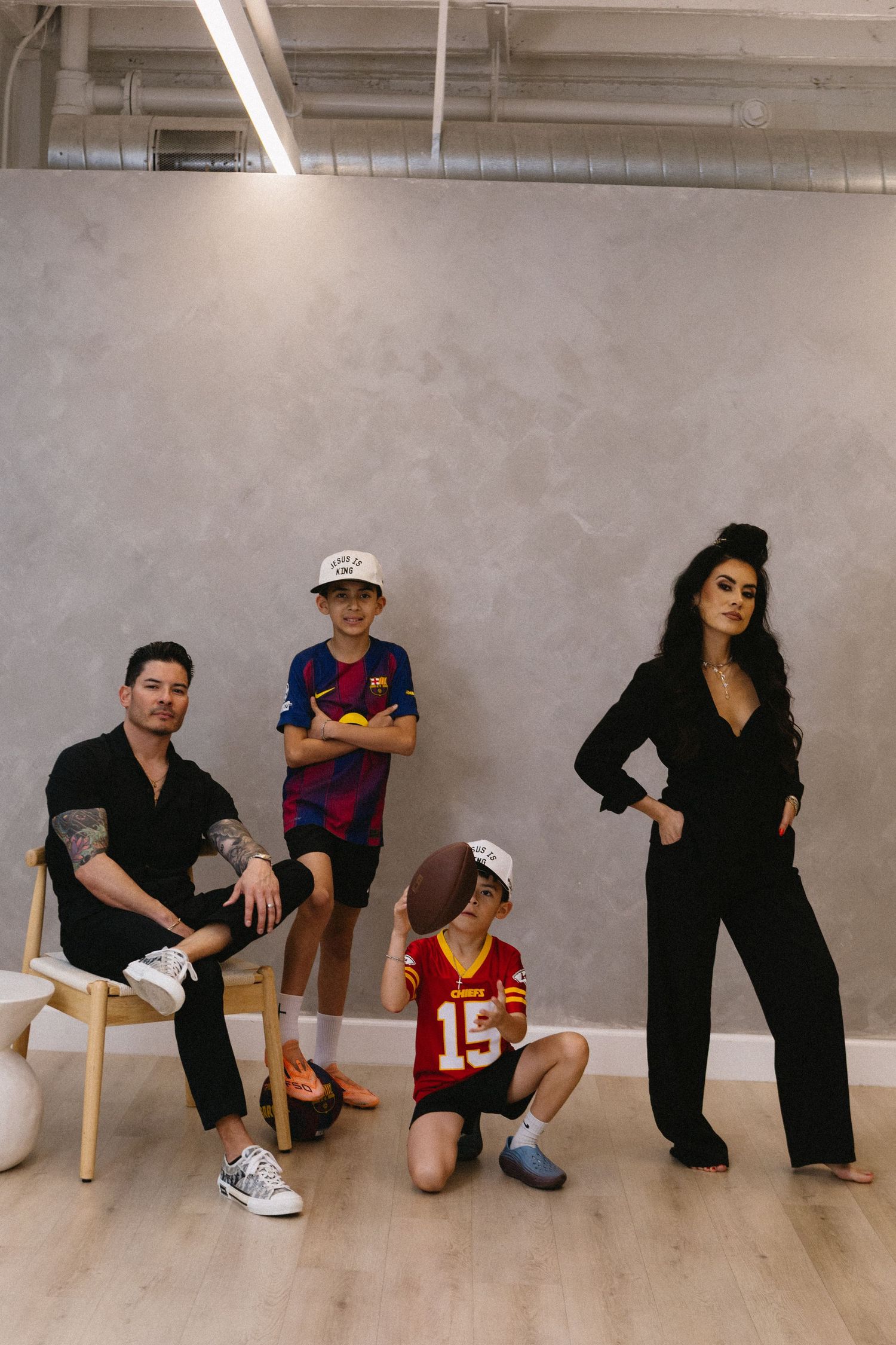 Family of four posing in modern studio, two boys in sports jerseys holding football, parents in stylish black outfits.