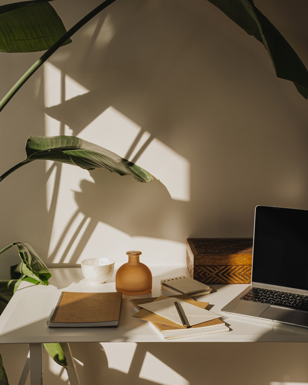 Minimalist workspace with laptop, vase and office supplies casting dramatic shadows in natural sunlight.