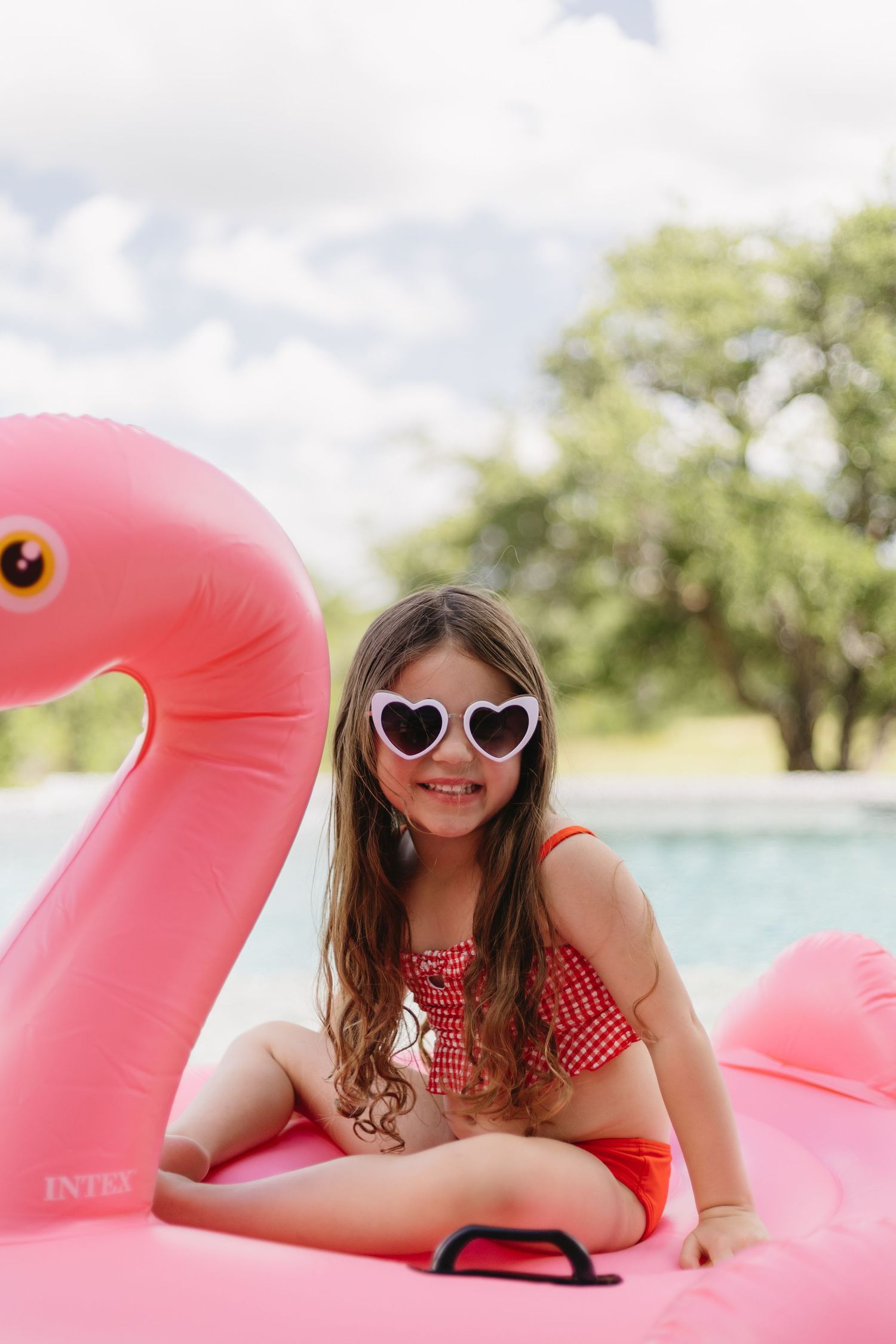 Person in heart-shaped sunglasses and red swimsuit relaxes on pink flamingo pool float on sunny summer day.