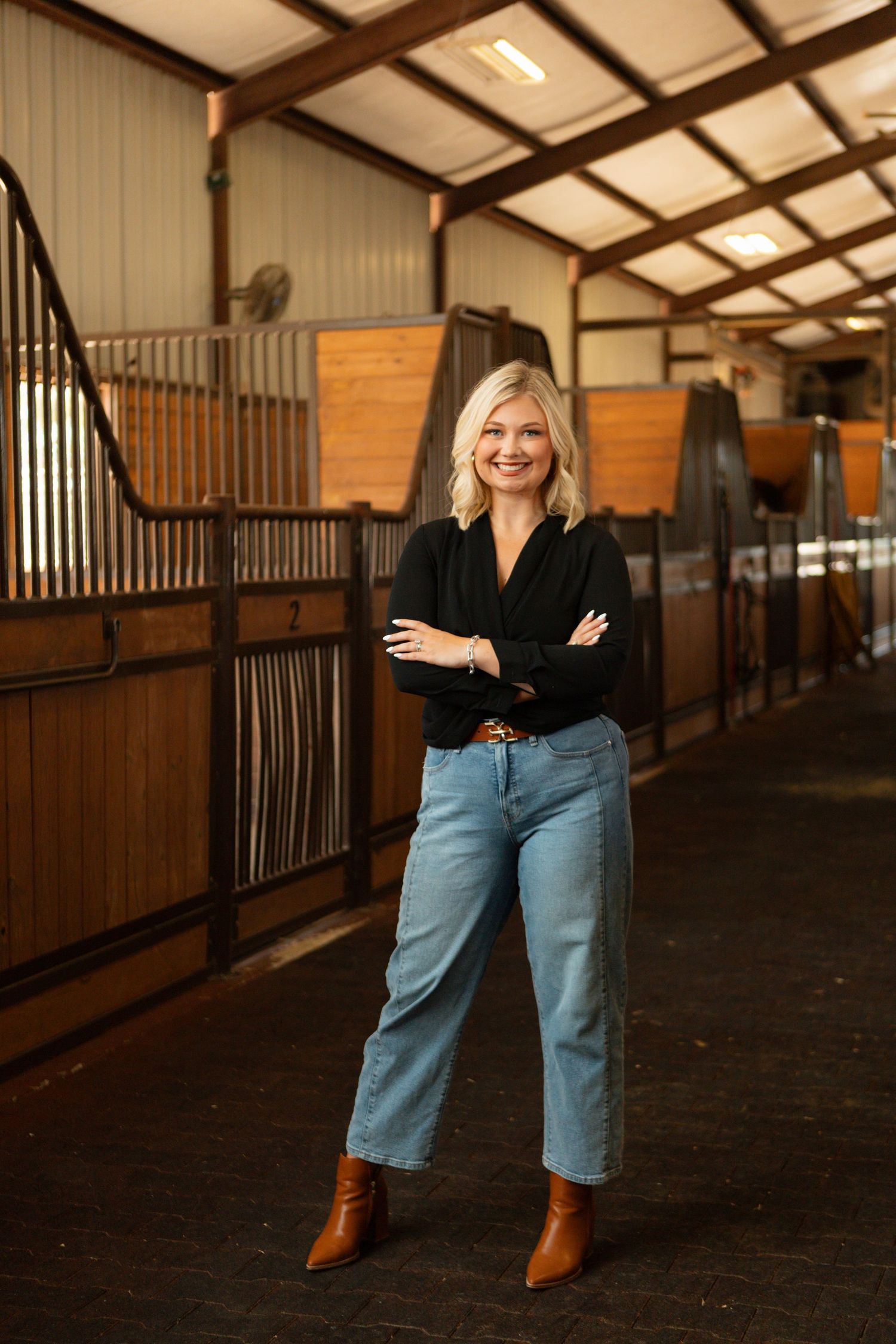 A person in a black top and light jeans stands confidently in a horse stable with wooden stalls and beams.