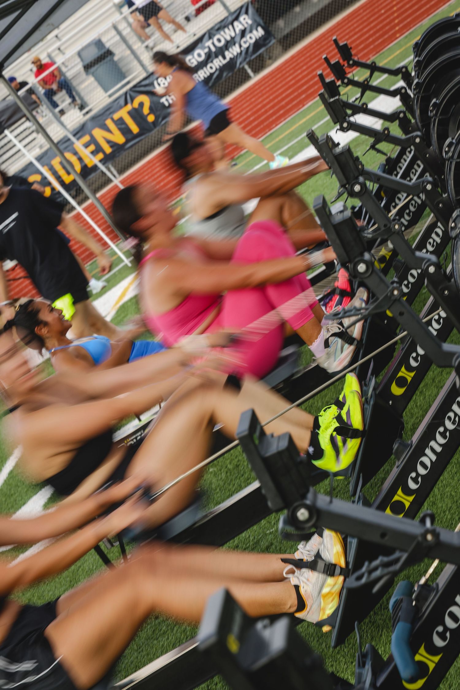 Athletes compete on Concept2 rowing machines indoors at a fitness event on artificial turf.