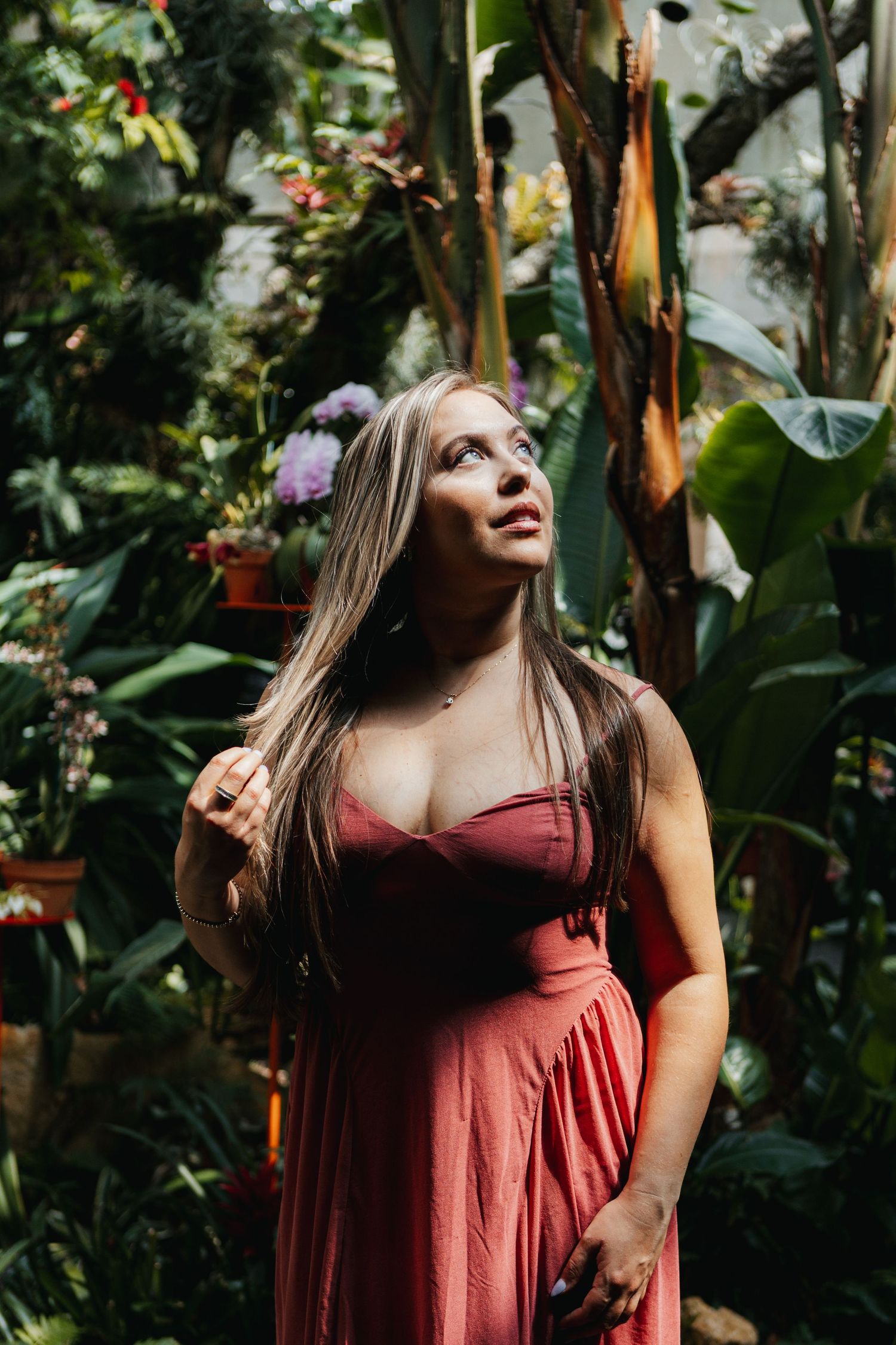 A person in a burgundy dress poses thoughtfully in a lush tropical garden setting with green foliage in the background.