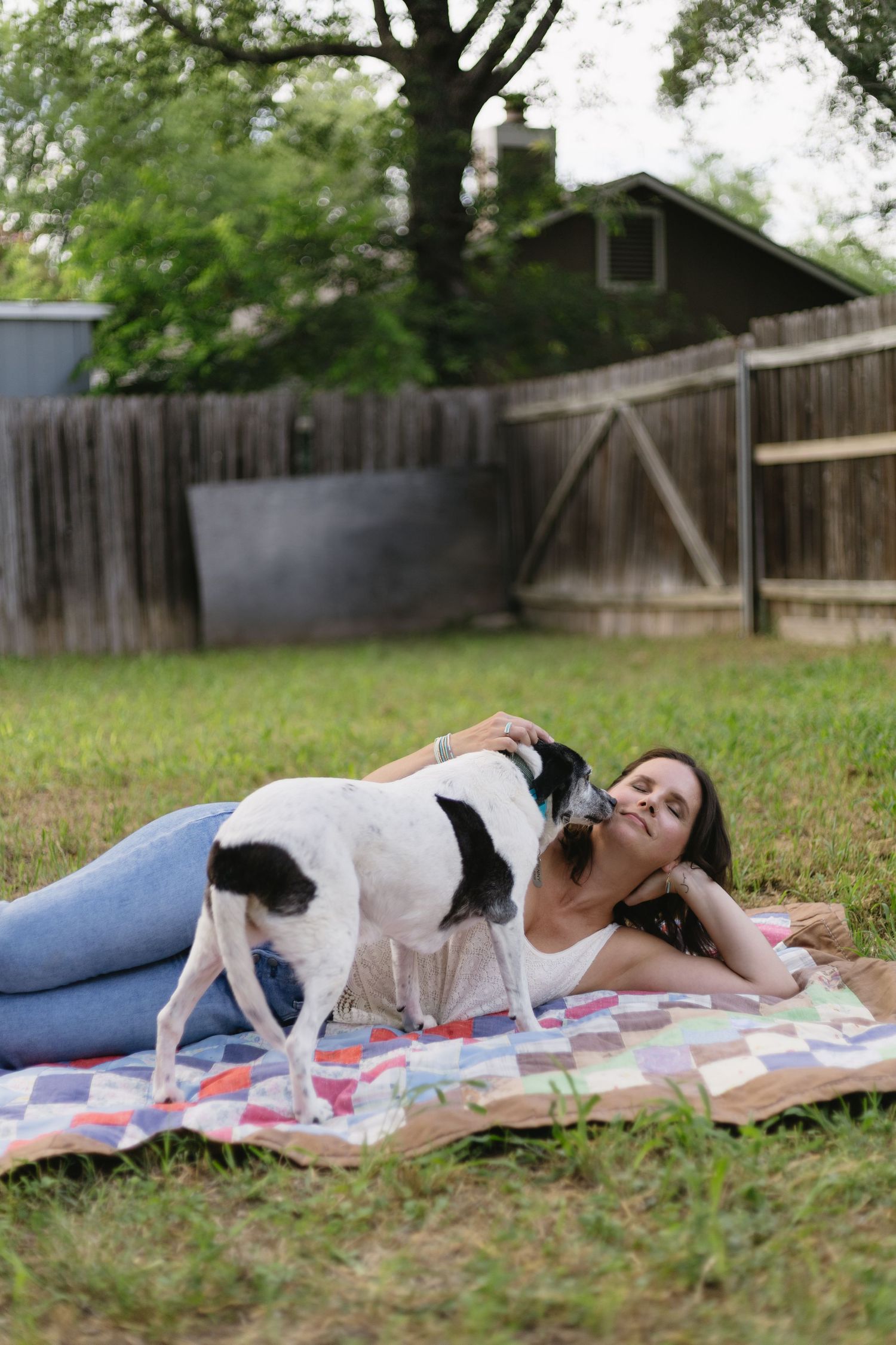 Black and white dog interacts playfully with someone laying on a picnic blanket in a backyard with wooden fence.