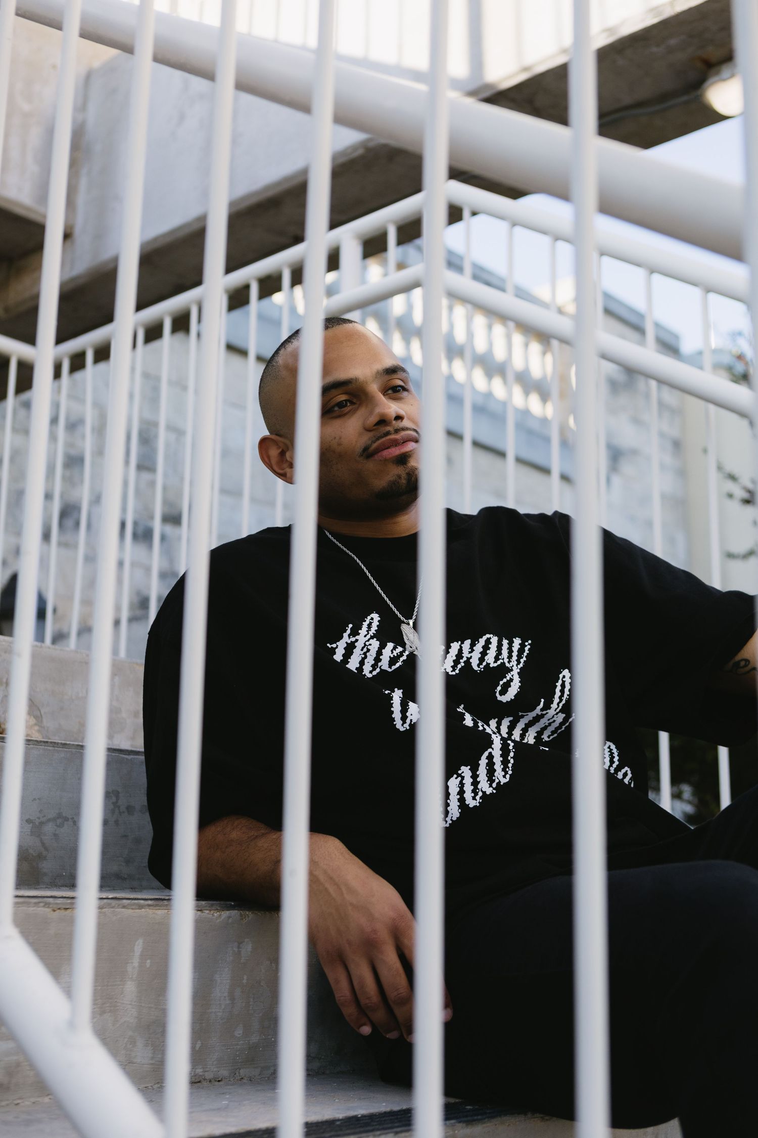 Bald man in black script-print sweatshirt sitting on steps behind white metal railings outdoors.
