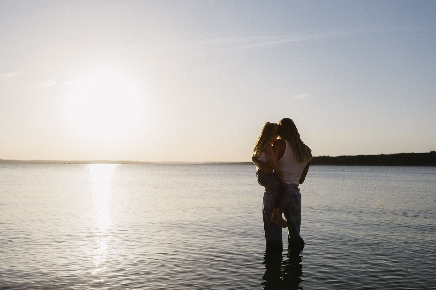 Silhouette of couple embracing in shallow water at sunset with golden light reflecting on calm lake.