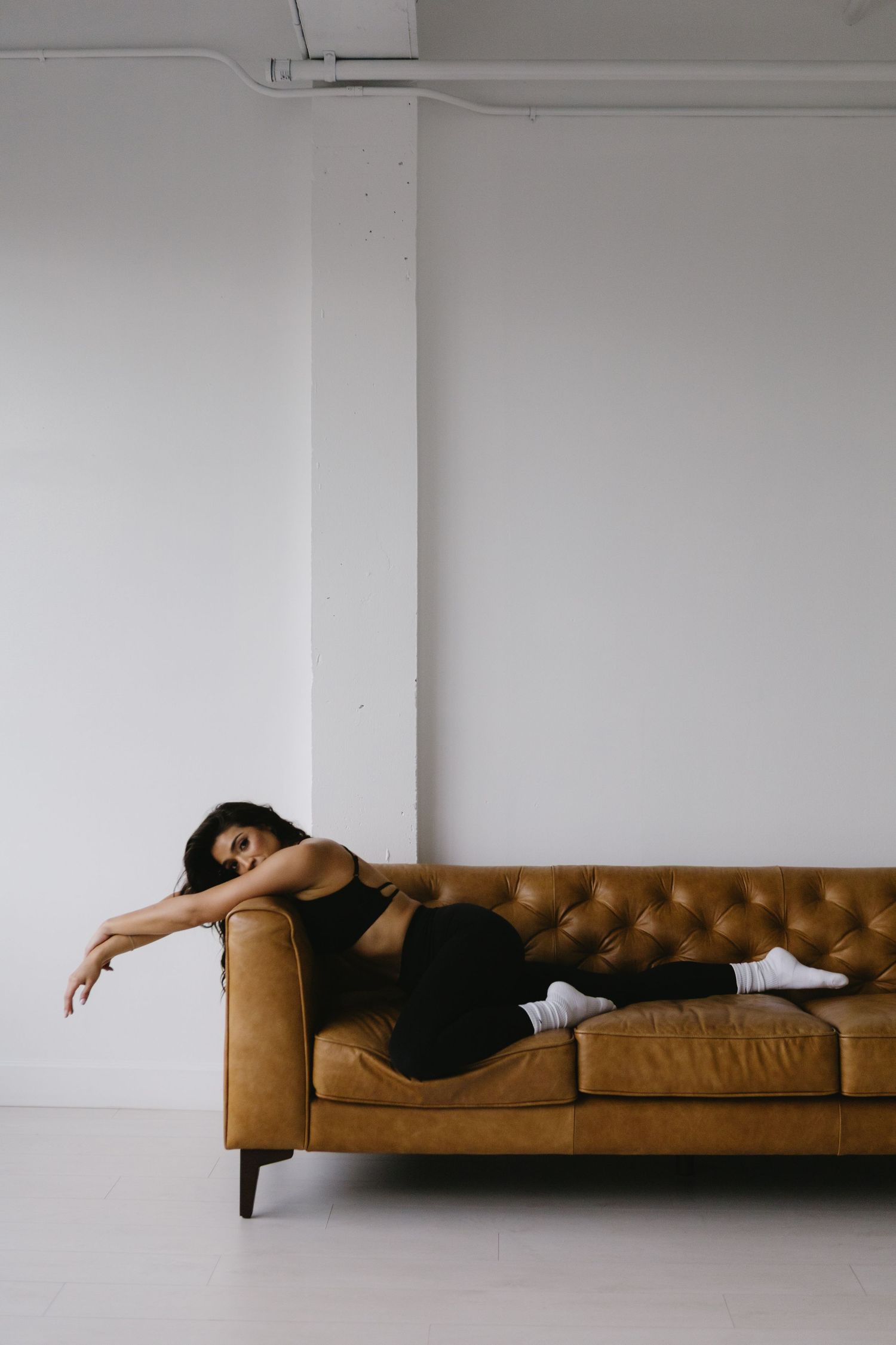 Woman in black athletic wear lounging on a tufted tan leather sofa against a white wall.