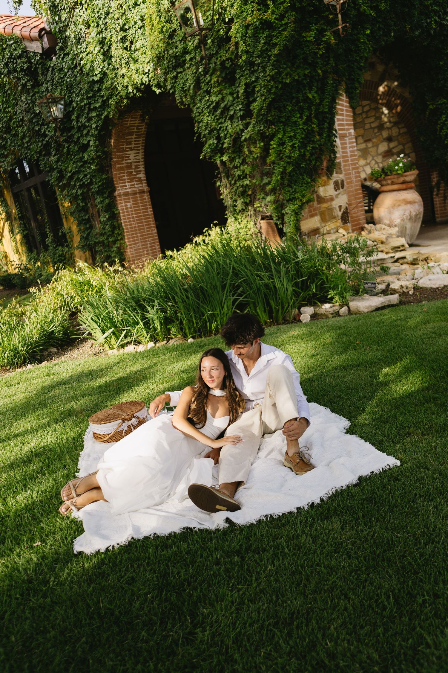Couple in white attire sitting on blanket in lush green garden near ivy-covered brick building.