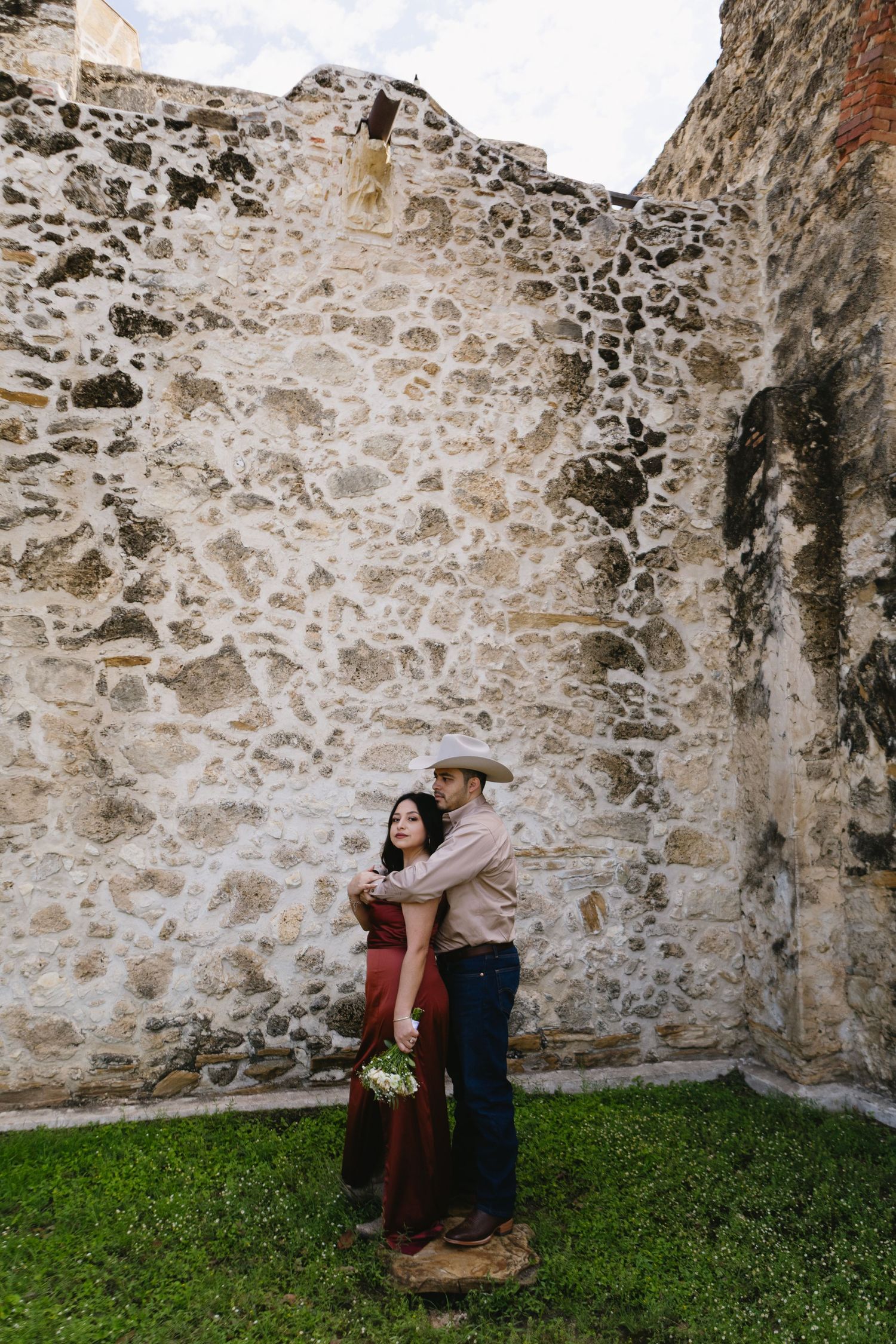 Couple embracing in front of a rustic stone wall, woman in red dress holding bouquet, man in cowboy hat and jeans.