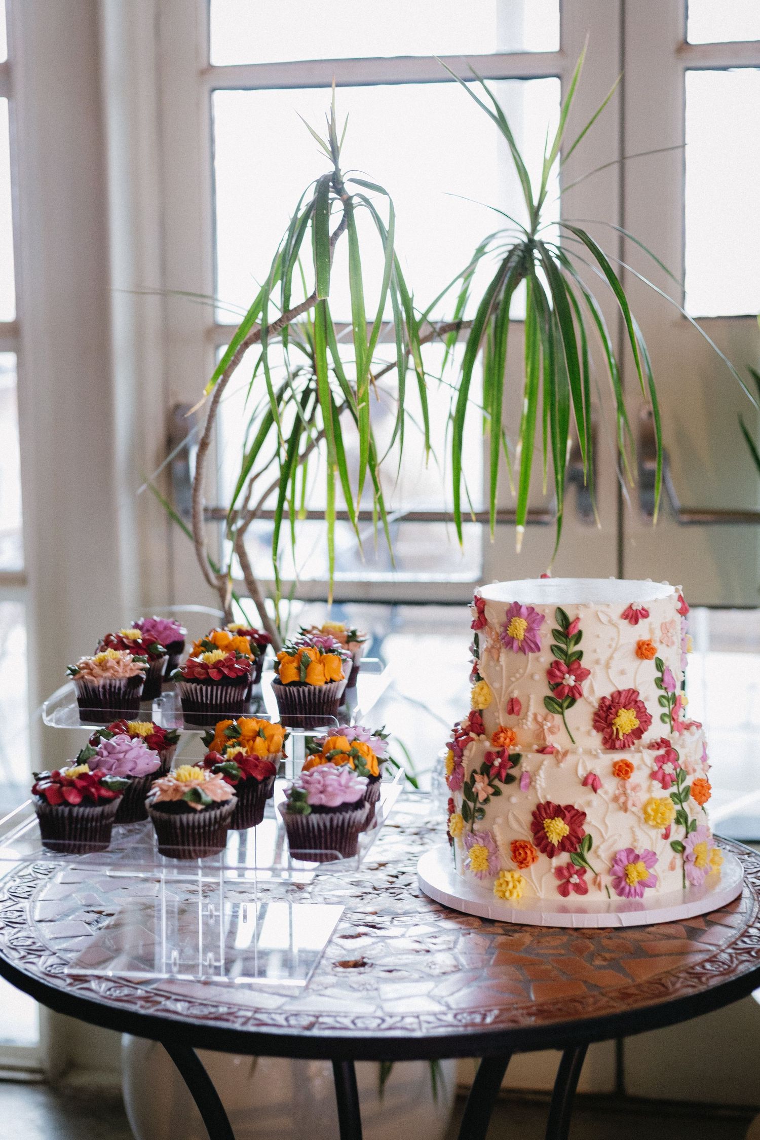 White two-tier wedding cake with colorful floral decorations beside cupcakes on a table with a plant backdrop.