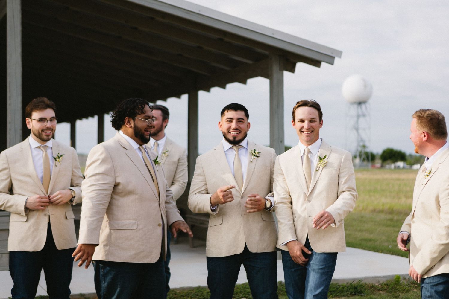 Groomsmen in tan suits with boutonnieres gather outdoors near a pavilion before a wedding ceremony.