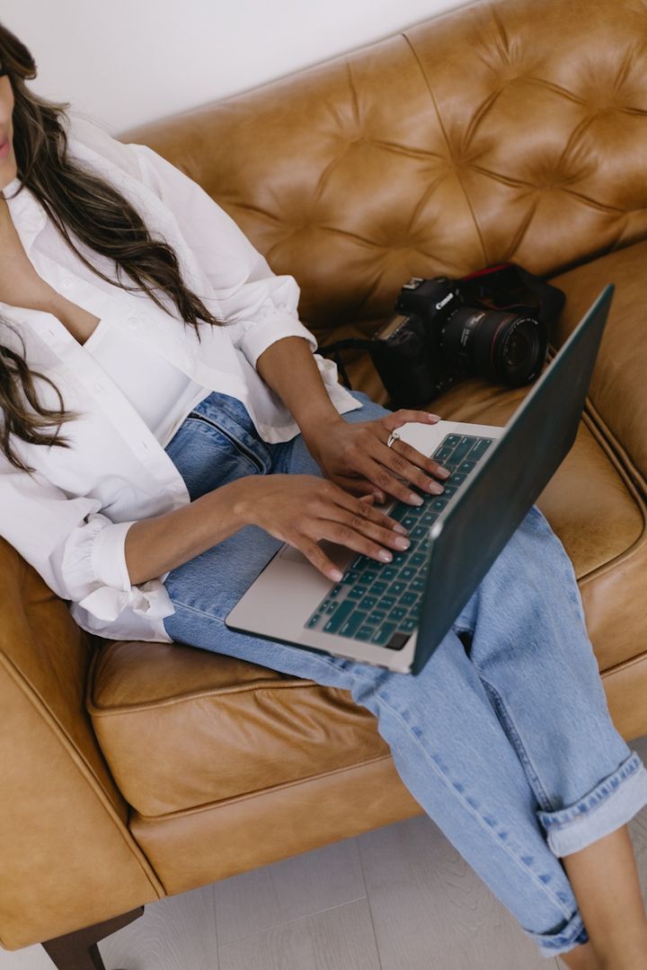 Person in white shirt and jeans working on laptop while relaxing on brown leather couch.