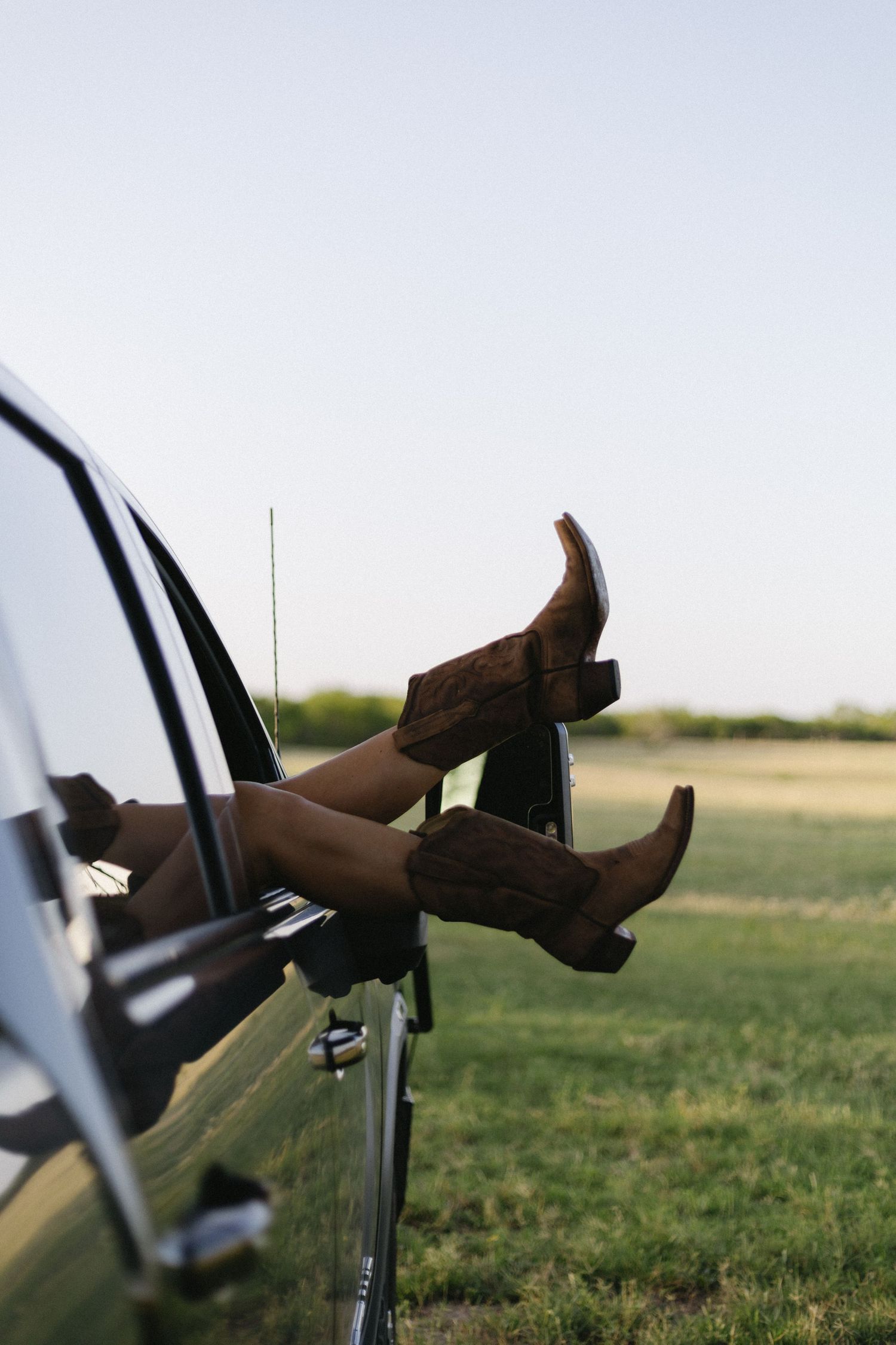 Brown cowboy boots sticking out of a car window parked in a grassy field during a sunny day.