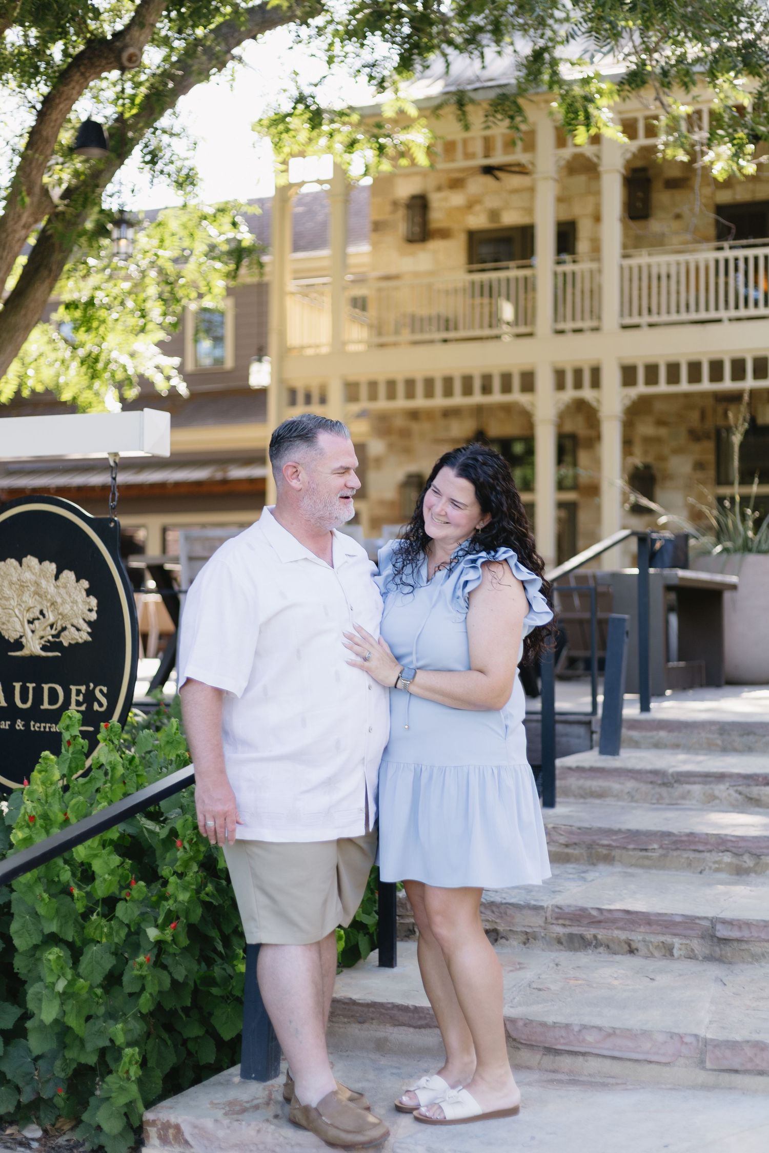 Couple smiling at each other outside Maude's restaurant on sunny day, woman in blue dress, man in white shirt.