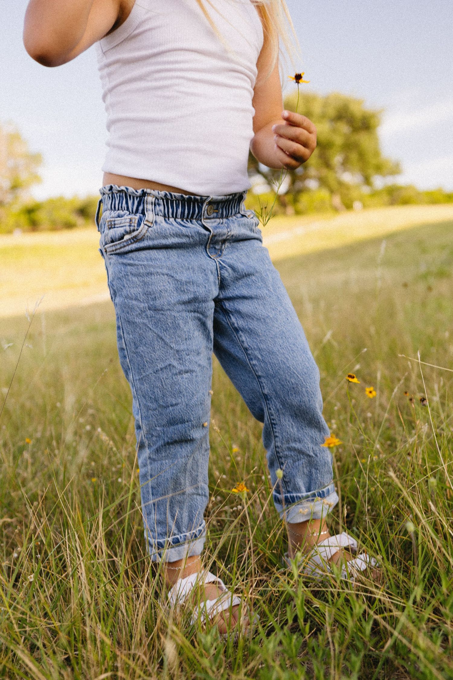 Child in blue jeans and white sneakers standing in a sunny green meadow holding a small yellow wildflower.