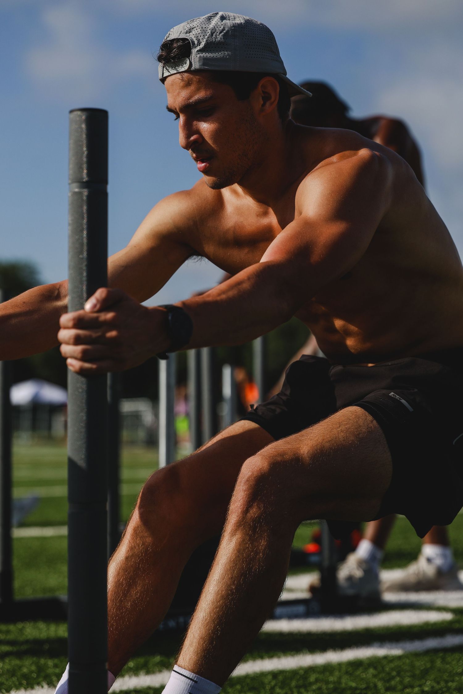 Muscular shirtless man wearing a cap pushes a weighted sled on a grass field during an outdoor fitness competition.