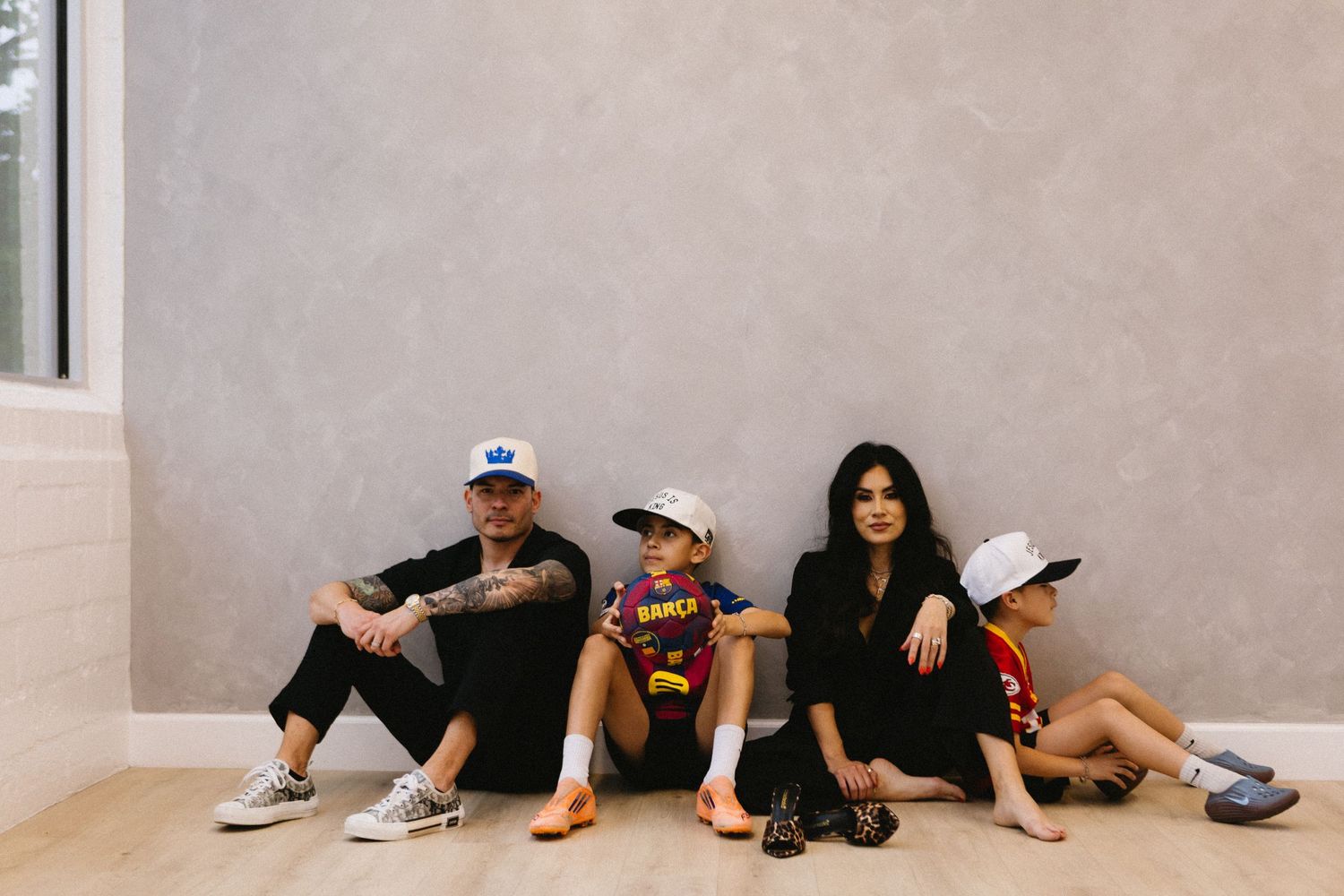 Family of four sitting casually on floor against gray wall, wearing hats and colorful outfits in a bright modern room.