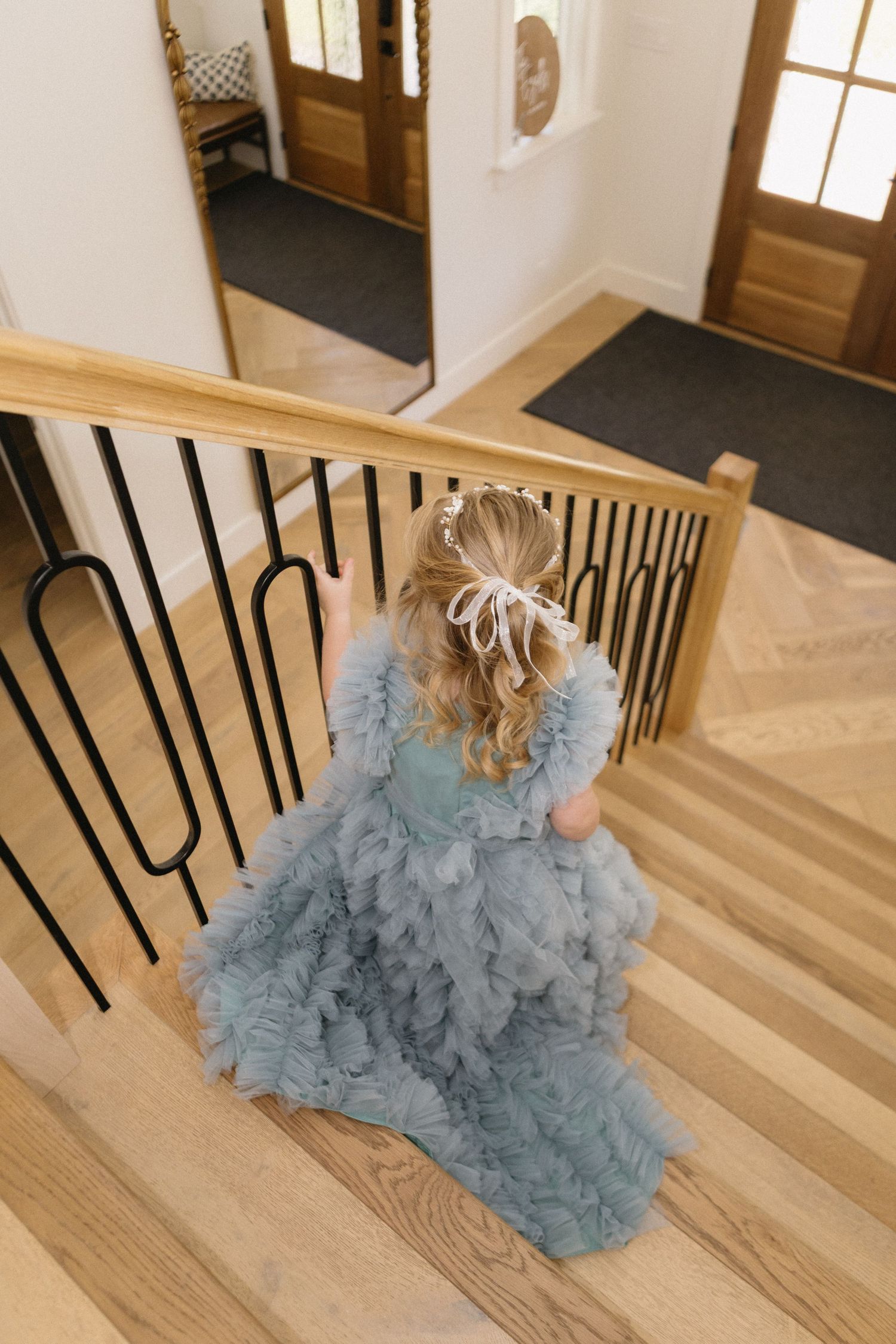 A young girl in a dusty blue ruffled tulle gown walks up wooden stairs, her curly blonde hair adorned with a bow.