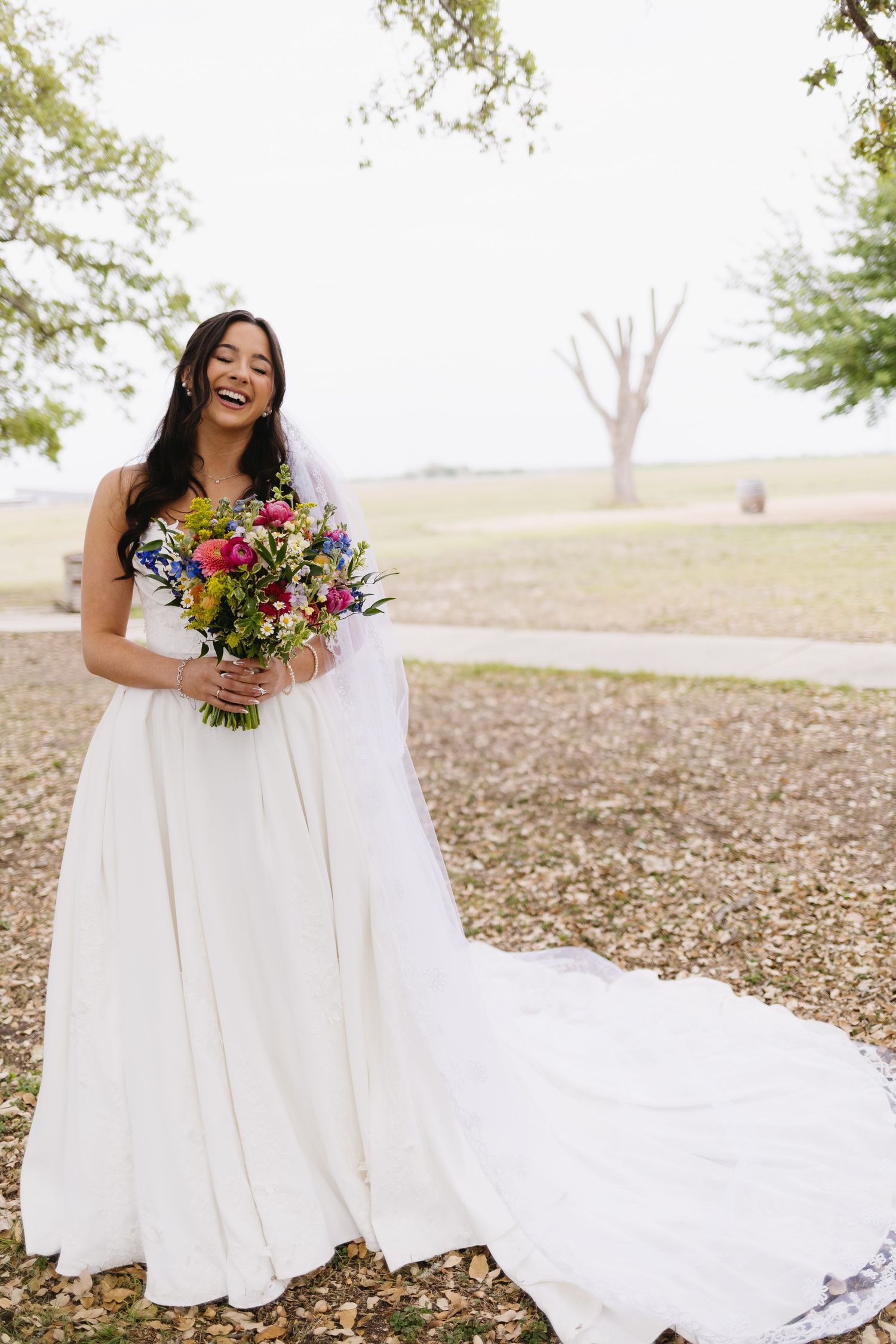 Smiling bride in white gown holds colorful bouquet outdoors on wedding day.